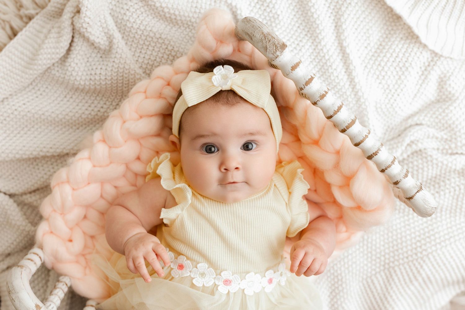 Adorable baby girl in yellow dress and bow headband lying on soft pink chunky knit blanket.