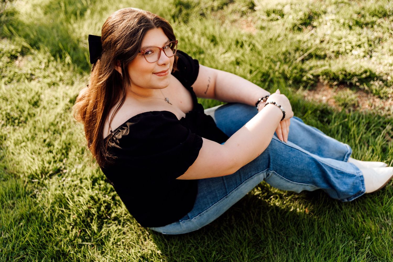 Young woman with glasses sitting on green grass in black top and jeans, posing for a portrait outdoors.