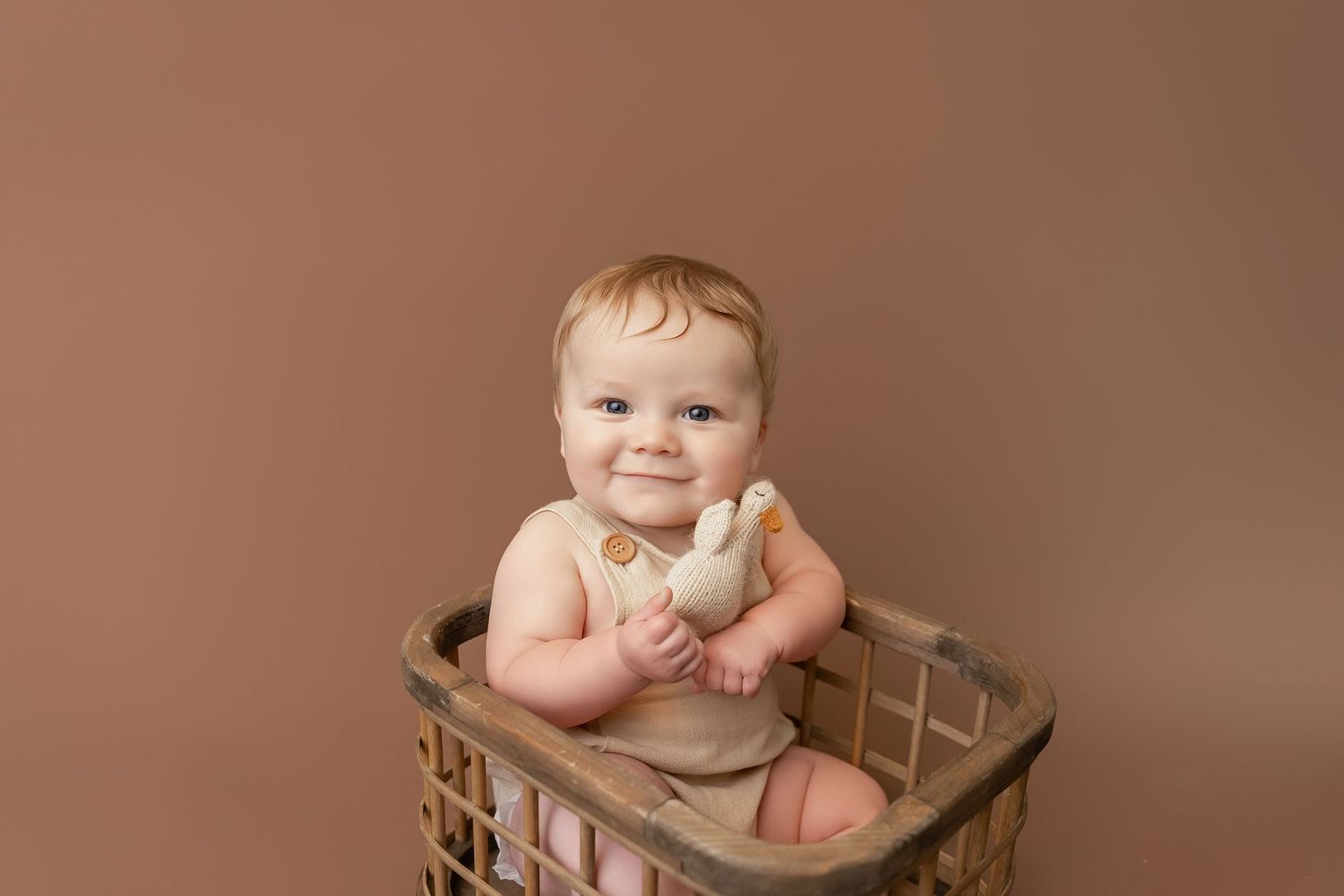 Smiling baby sitting in a rustic wooden basket holding a stuffed duck toy against a warm brown background.