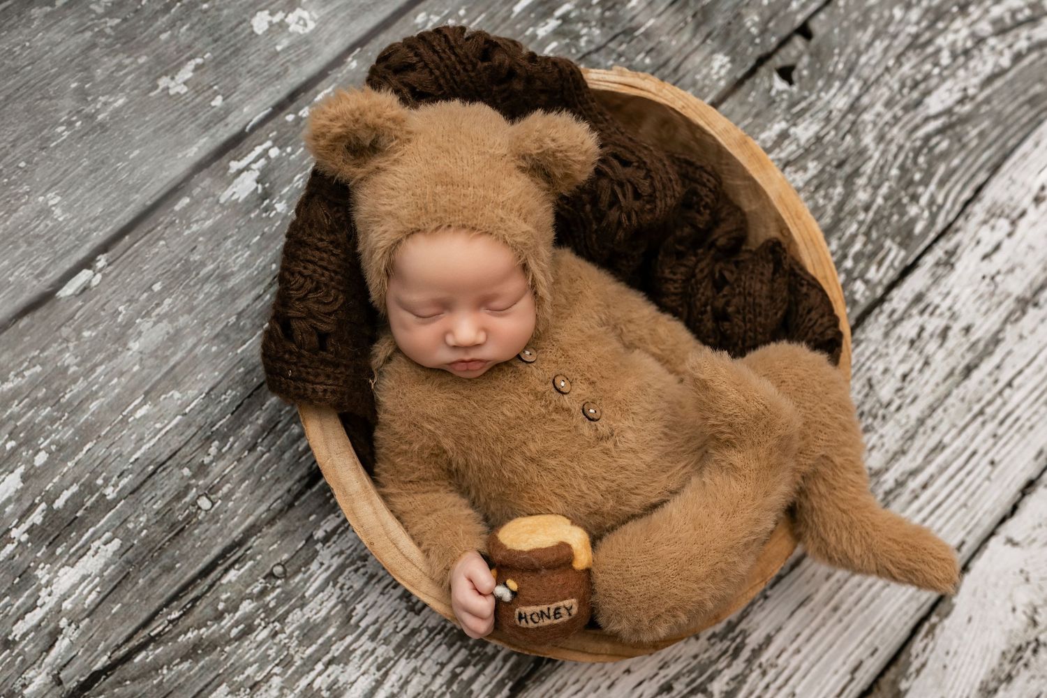Newborn baby dressed in brown bear fuzzy outfit and bonnet sleeping in wooden bowl holding honey pot.