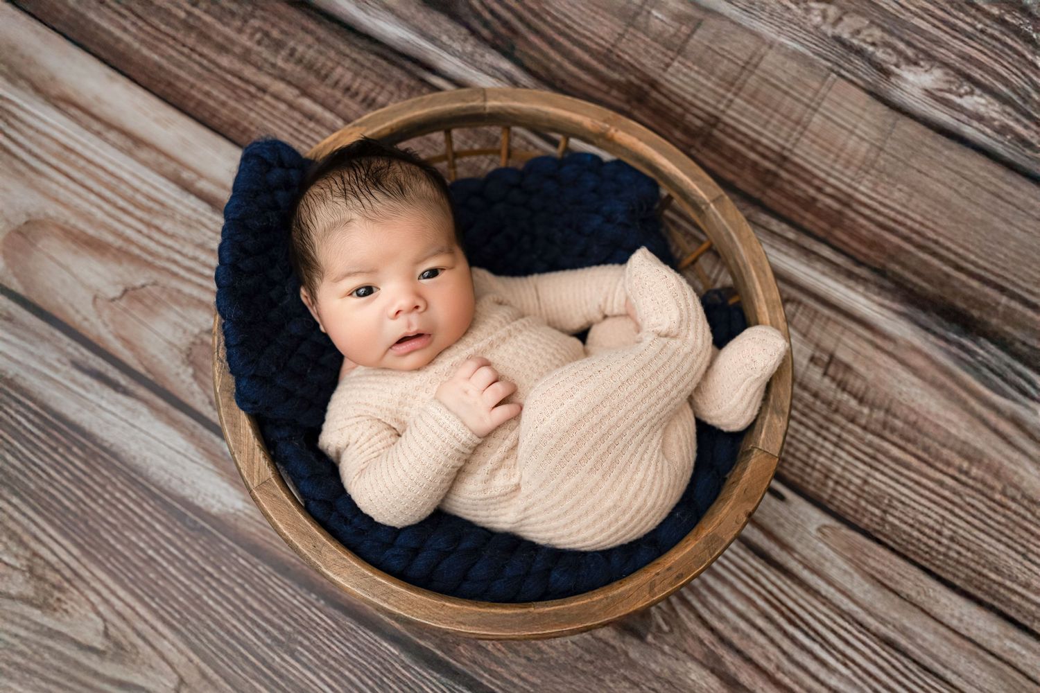 Newborn baby in pink knit outfit sleeping in a wicker basket with navy blue blanket on wood floor.
