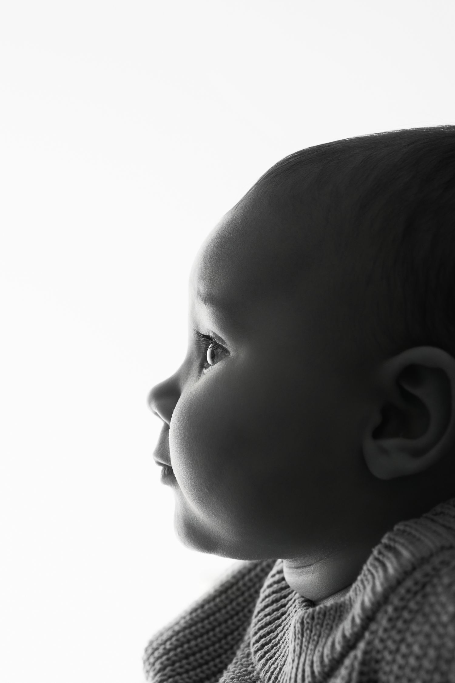 Black and white side profile portrait of a baby in a knit sweater, softly backlit against a bright white background.