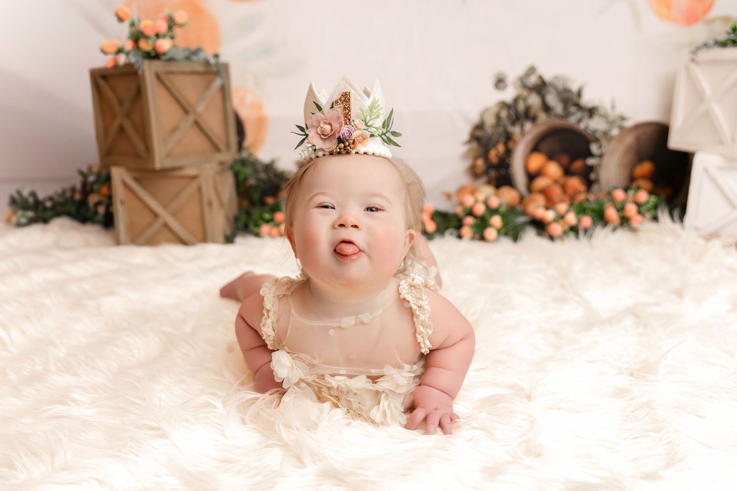 Baby wearing a floral crown and lace outfit lies on a fluffy white rug with tongue out, surrounded by wooden crates and orange flowers.