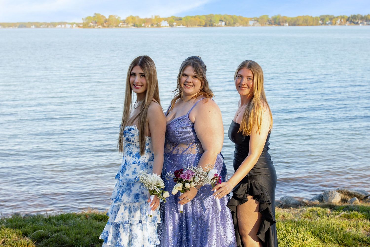 Three young women in formal prom dresses pose together by a lakeside on a sunny day.