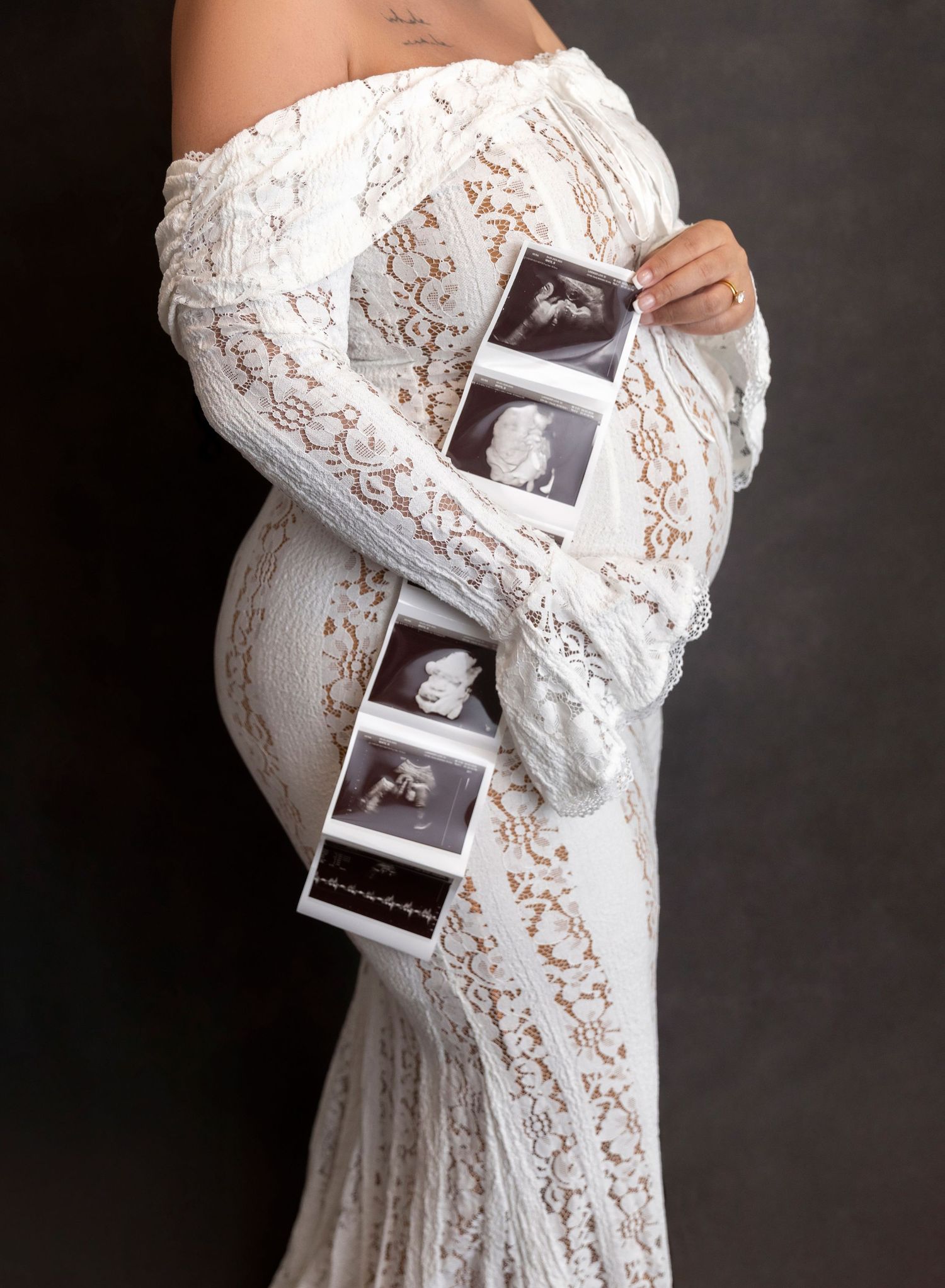 Pregnant woman in white lace off-shoulder gown holding ultrasound sonogram strip, maternity photography portrait.