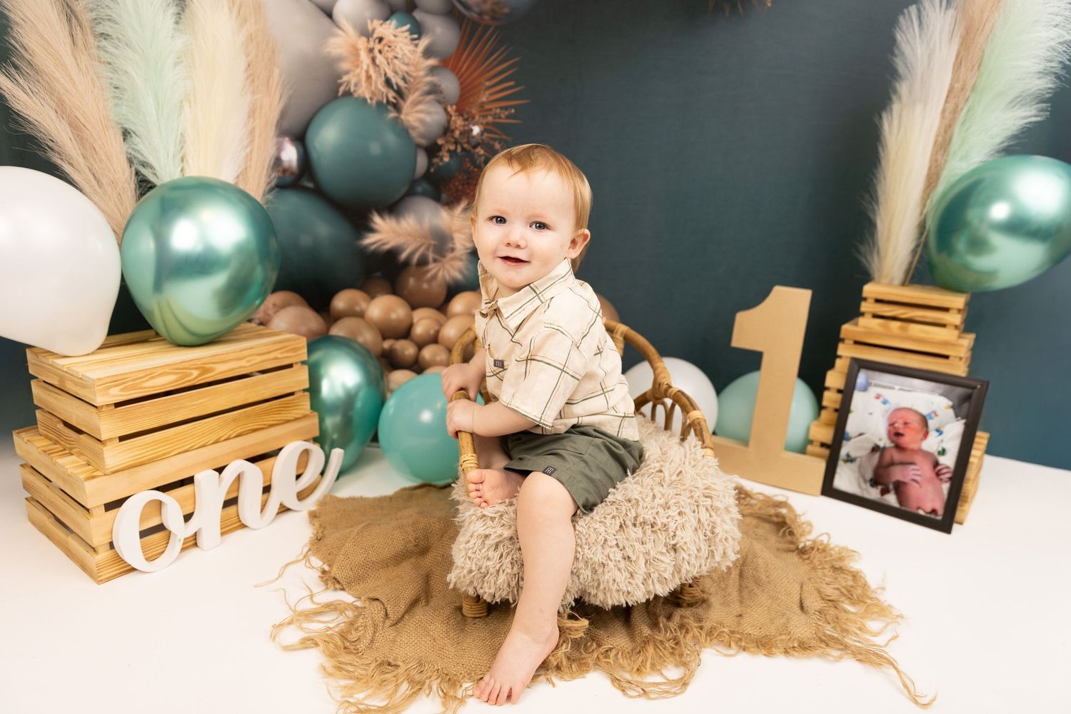 Baby sits on brown blanket surrounded by teal balloons and wooden crates for first birthday photo session.
