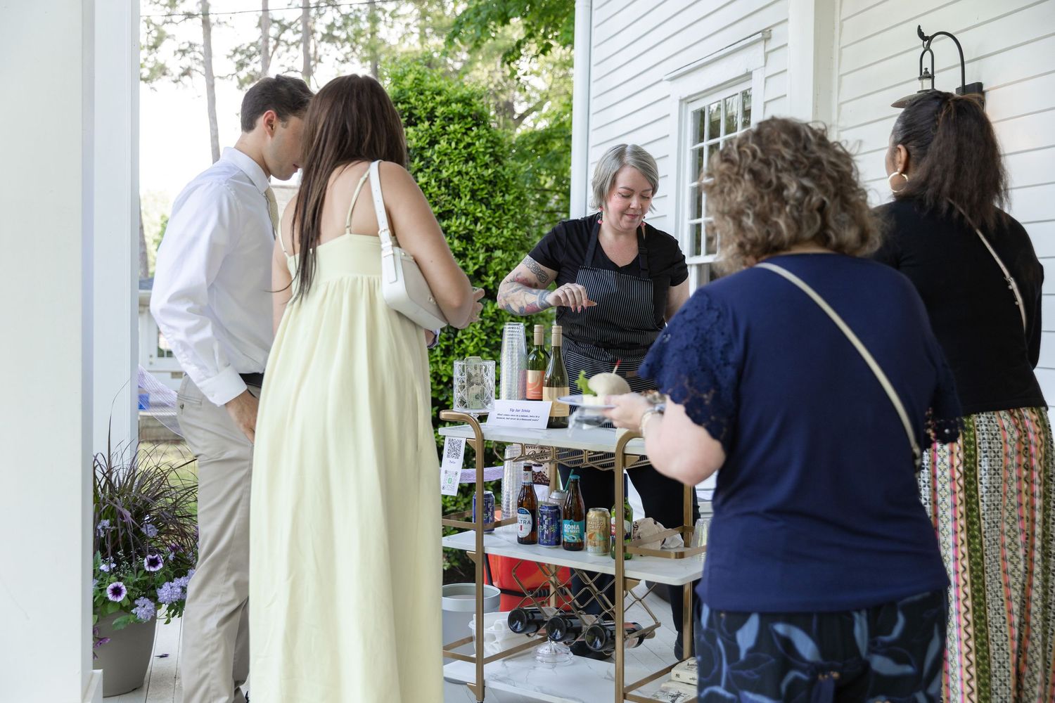 Group of people socializing on a porch around a drink cart at an outdoor gathering.
