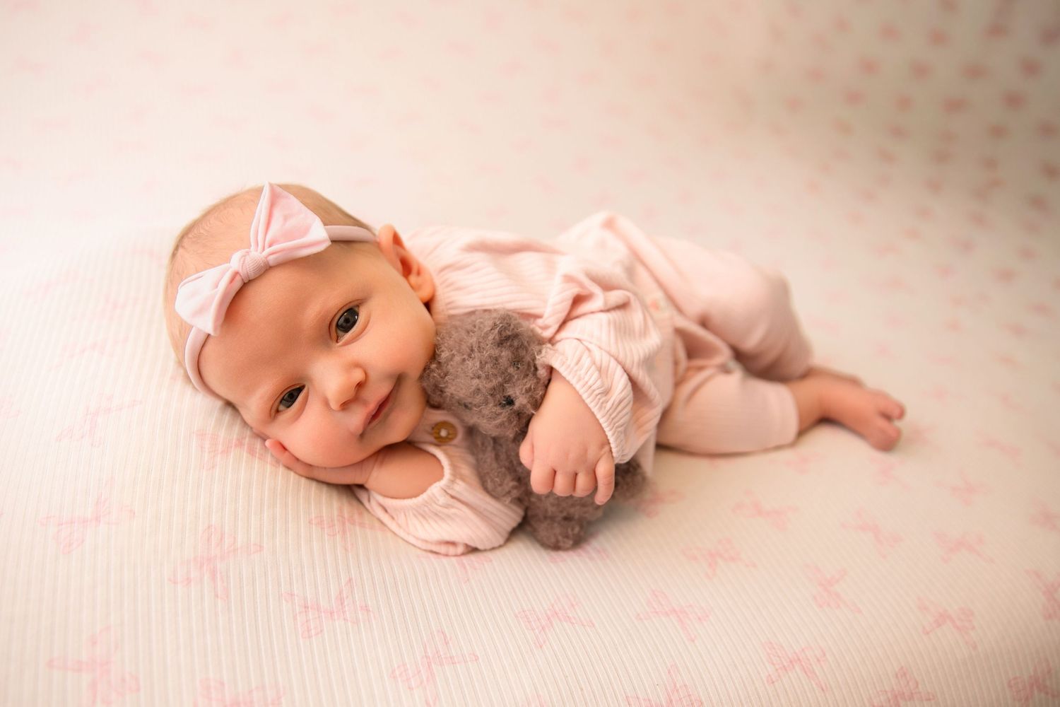 A baby in pink clothing lies on a soft white blanket while cuddling with a plush teddy bear.