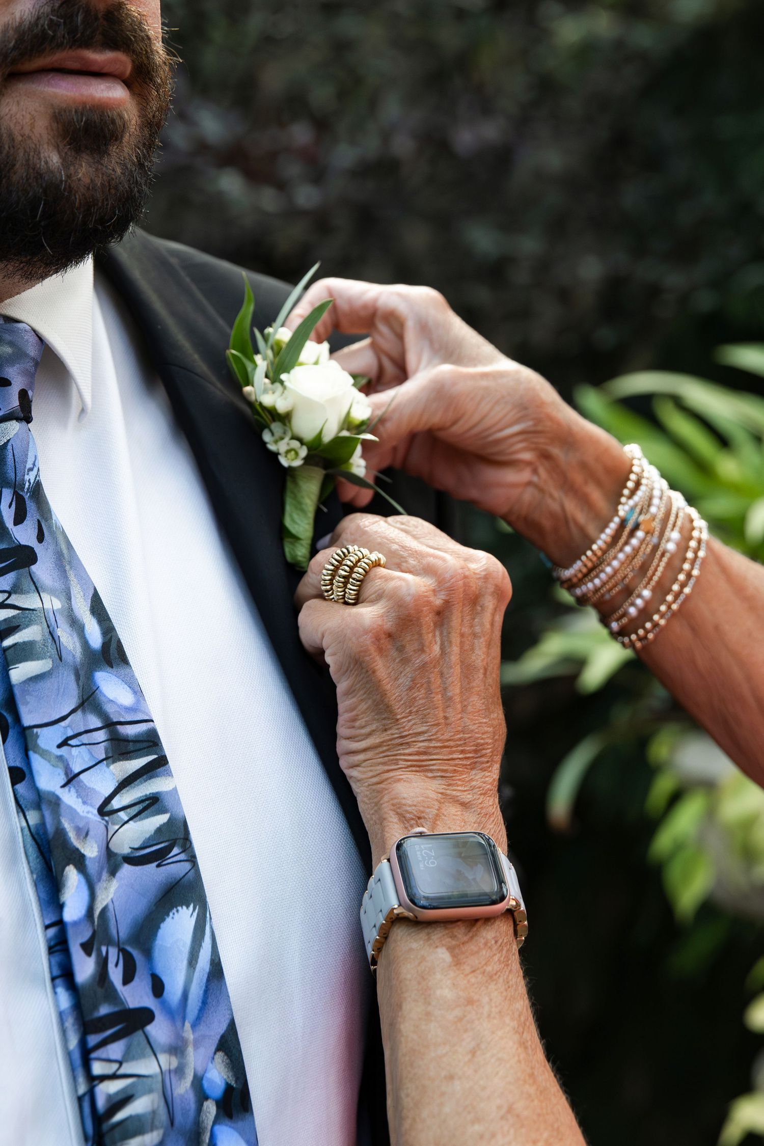 Elderly woman pinning a white rose boutonniere on a bearded groom wearing a blue floral tie at a wedding.
