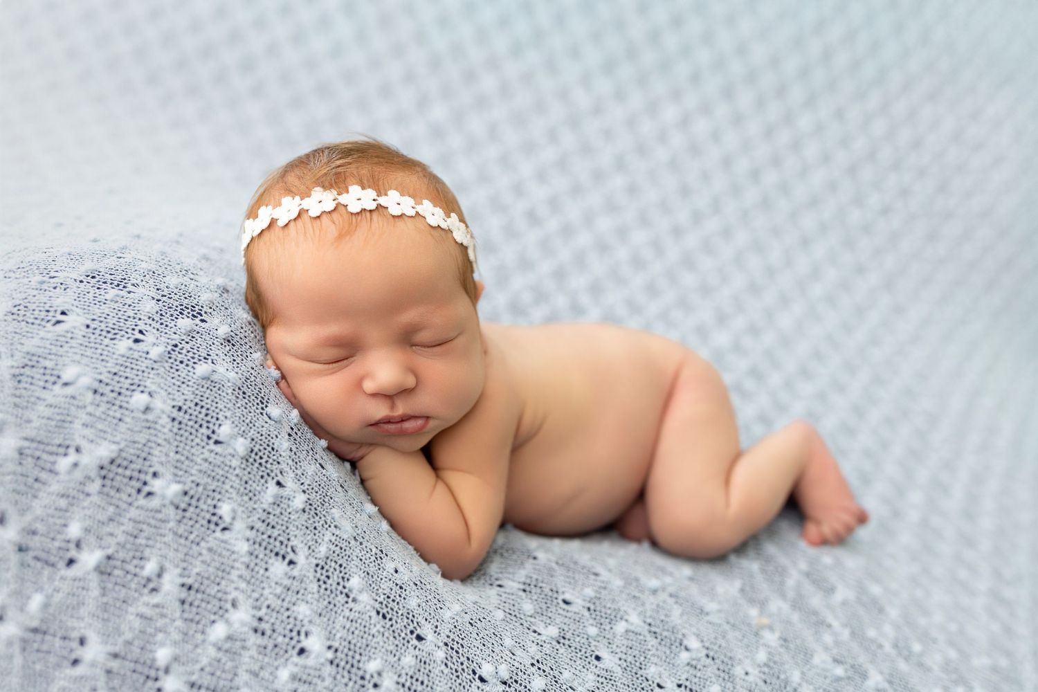 Newborn baby sleeping peacefully on blue fabric wearing a delicate white floral headband.