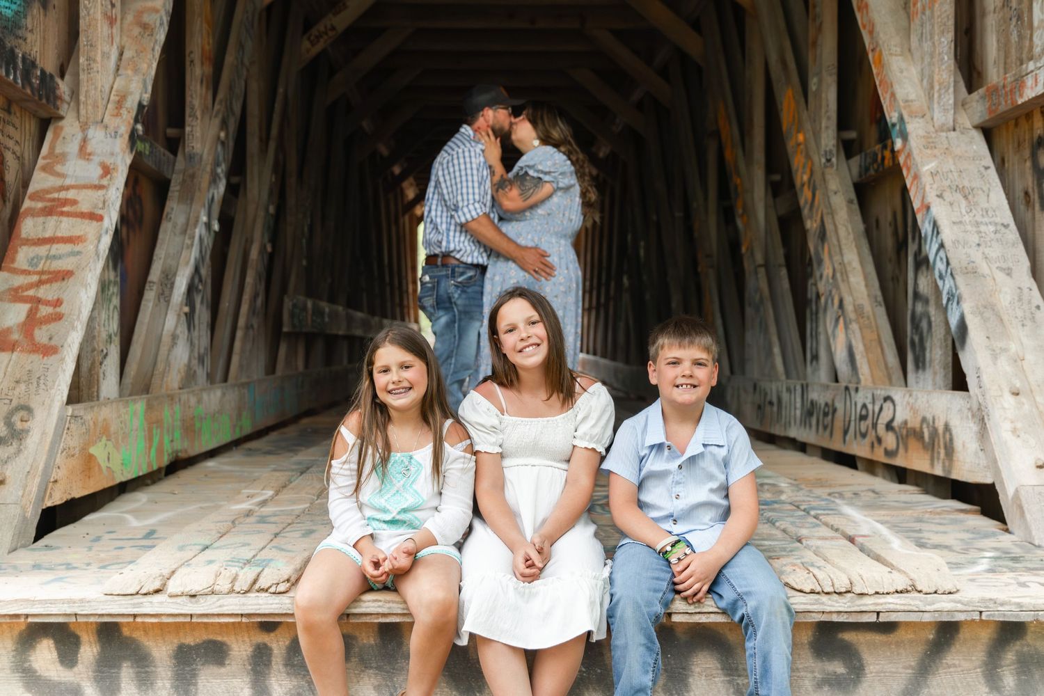 Family portrait inside a rustic covered bridge with three children in front and parents embracing behind them.