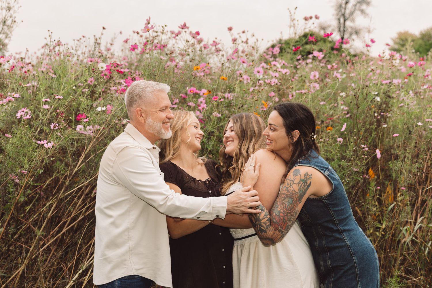 A gray-haired man and three women embrace in a joyful family portrait among blooming pink wildflowers in a sunlit field.