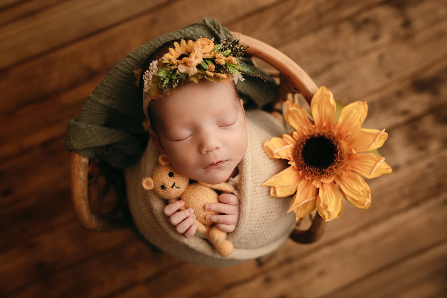 Sleeping newborn baby in a floral headband cradling a tiny teddy bear in a wooden bowl beside a sunflower on a rustic wood floor.