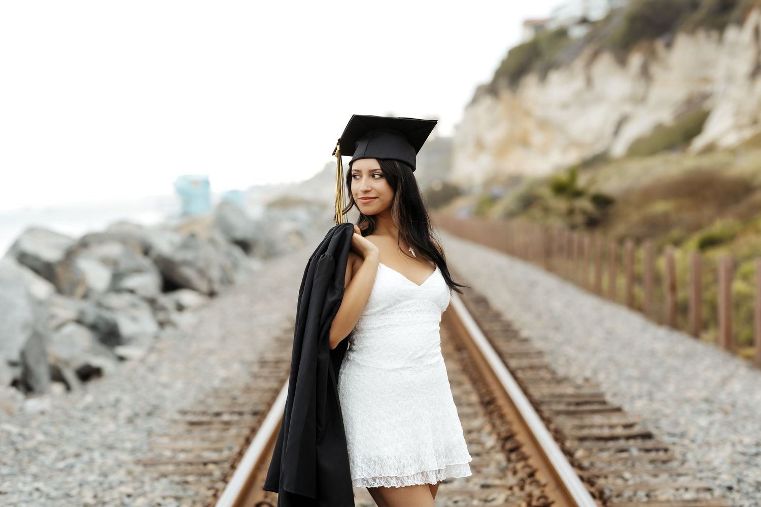 A confident graduating senior holding her gown over her shoulder wearing her cap looking out to the side in San Clemente, CA