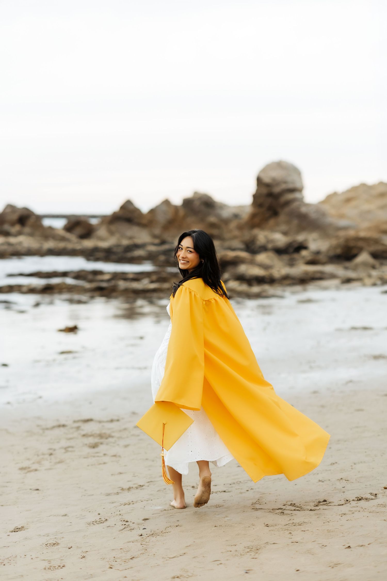 High school graduate in cap and gown walking on the beach in Orange County
