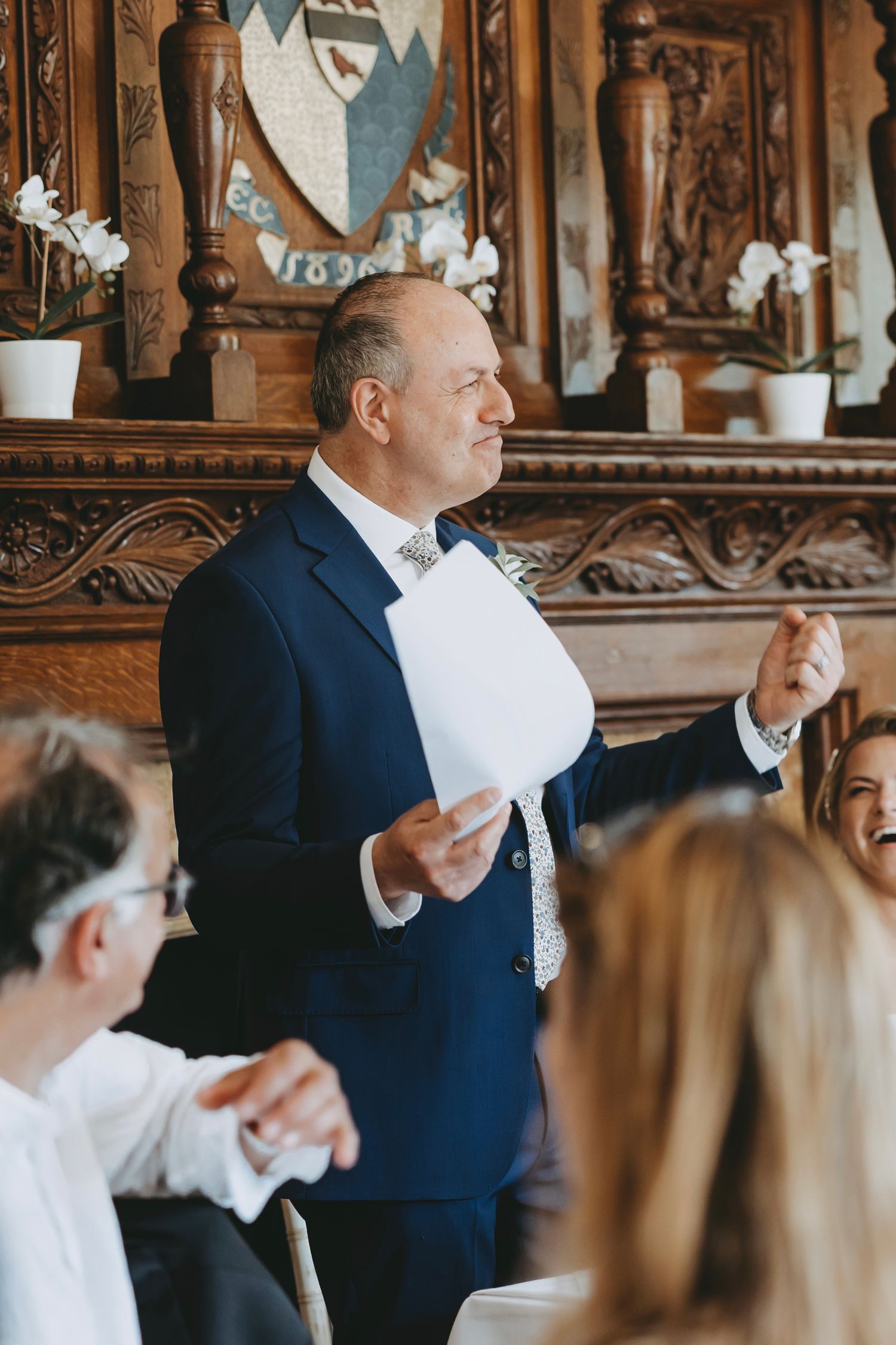 Wedding guests and speakers interact during a reception in an ornate wood-paneled venue.