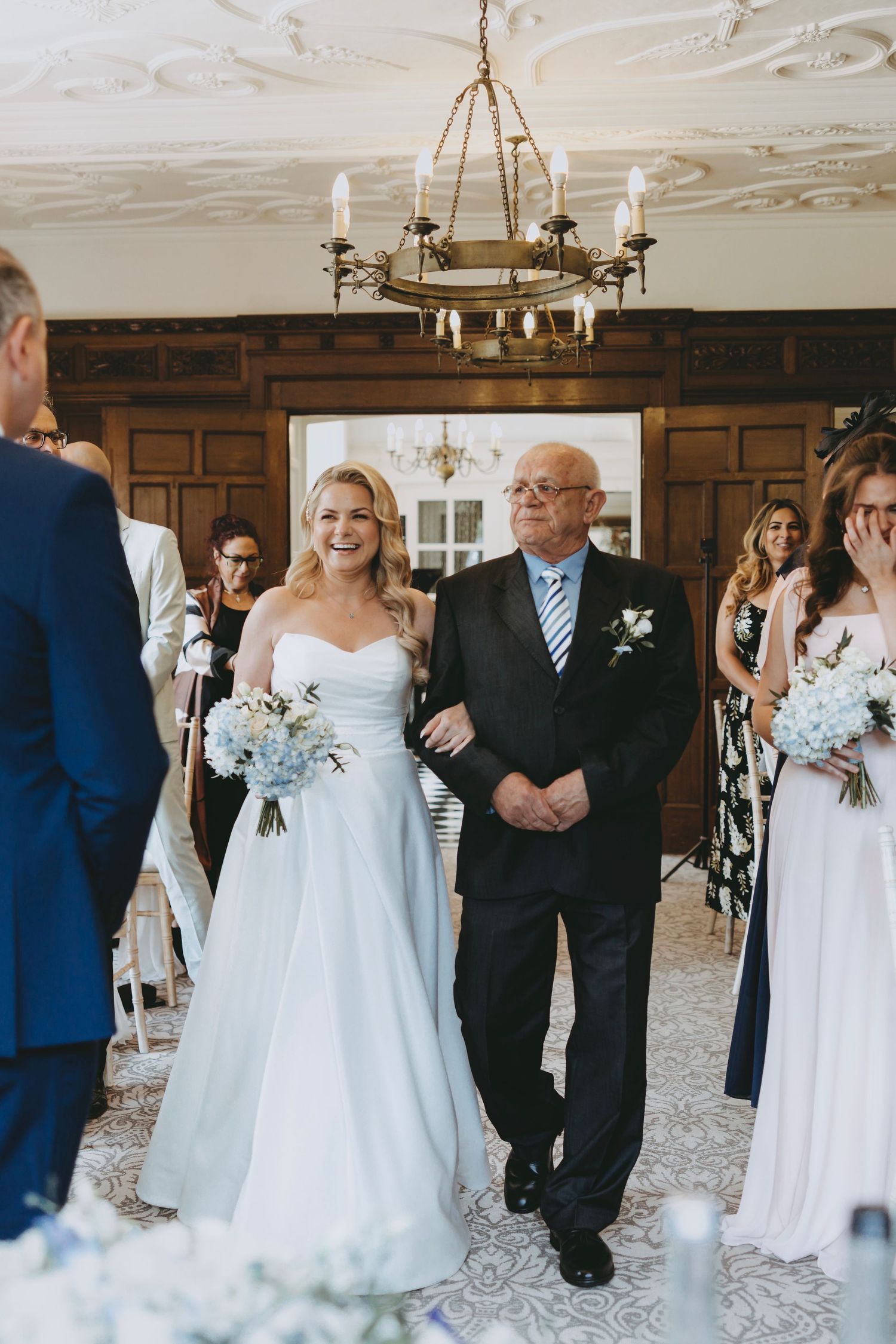 Wedding party members in formal attire standing beneath ornate chandeliers in wood-paneled room.