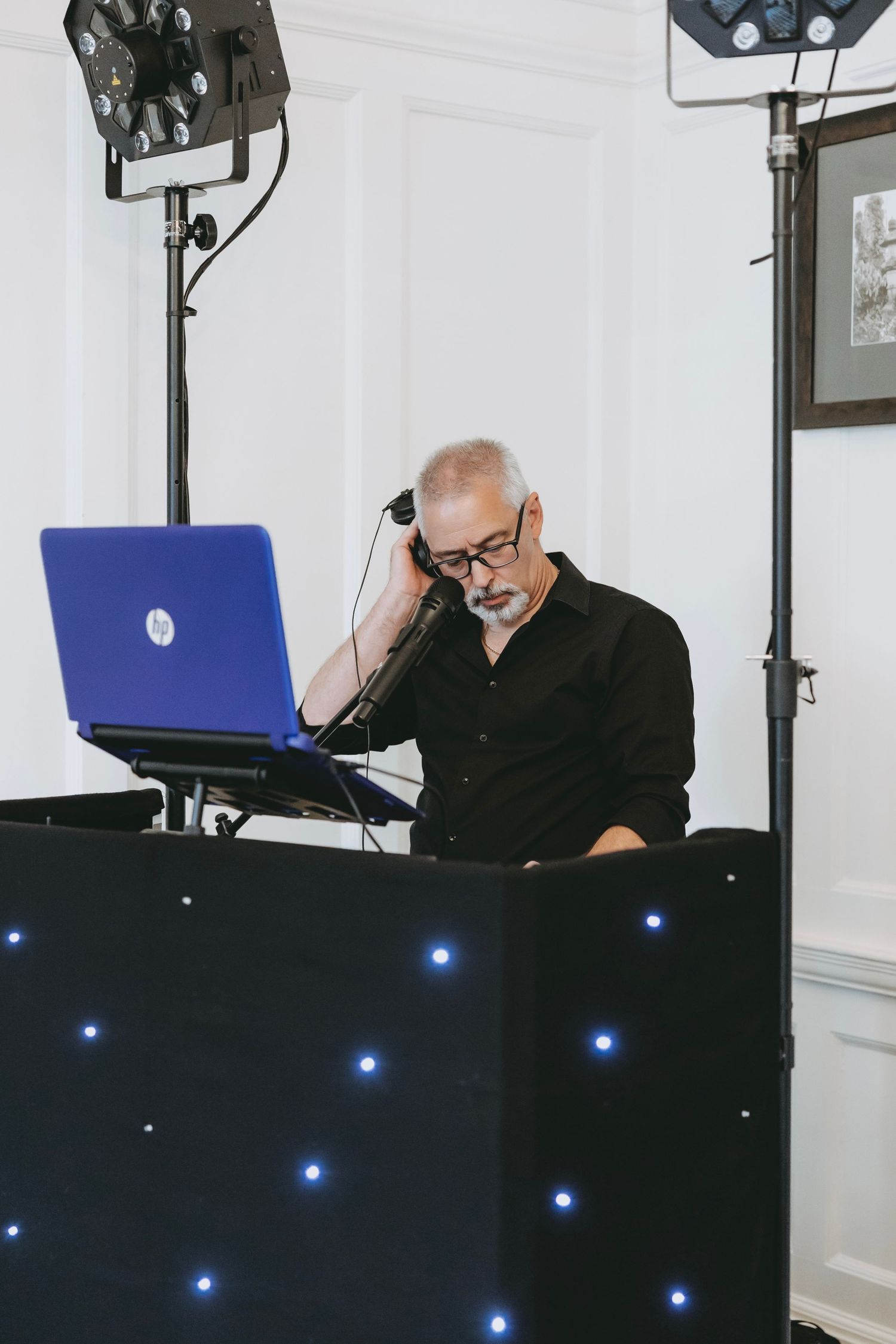 DJ wearing headphones operates equipment at a wedding reception behind an illuminated booth.