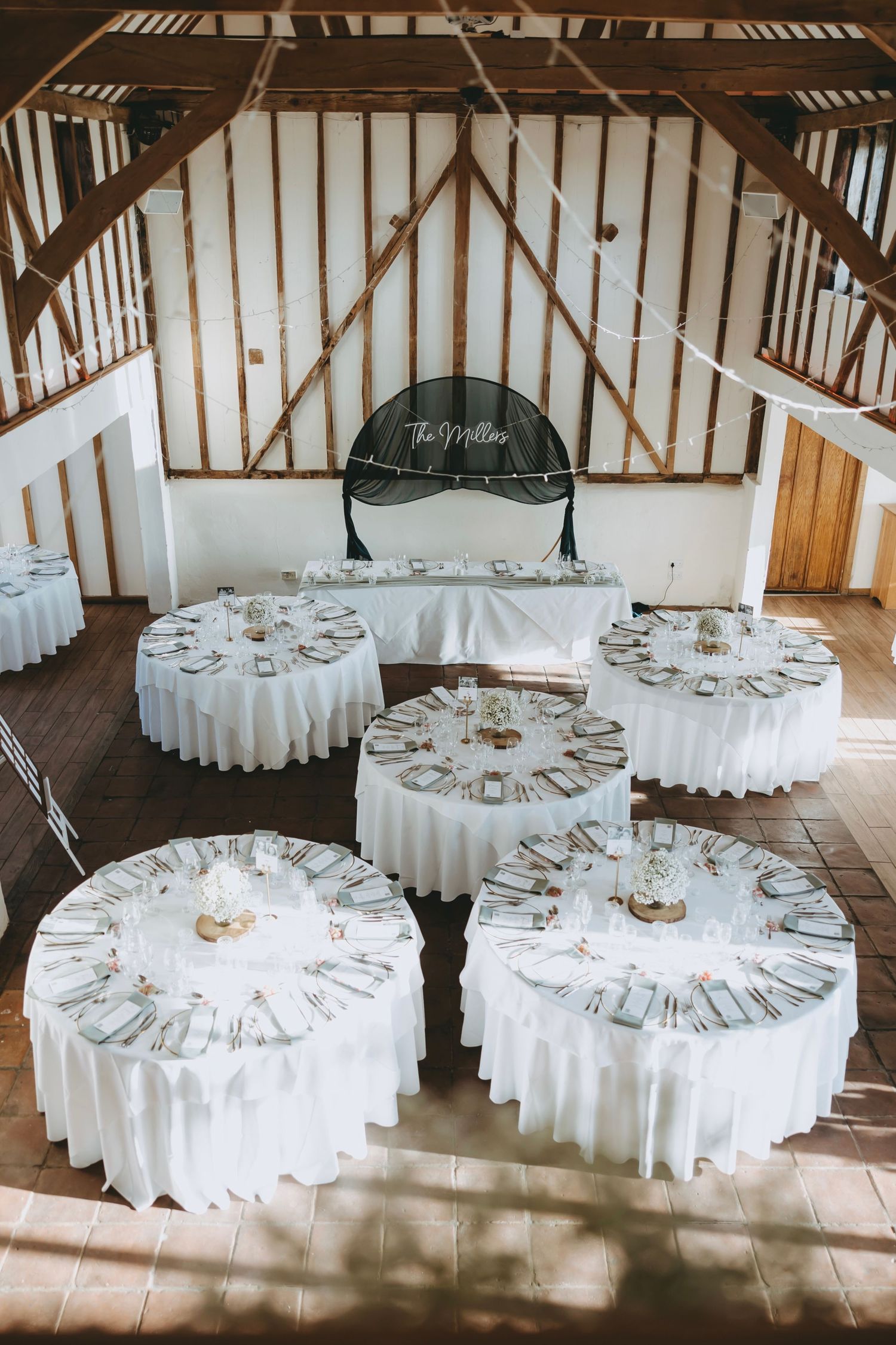 Wedding reception venue with white round tables set up under exposed wooden beams at Coltsfoot Retreat, Hertfordshire.