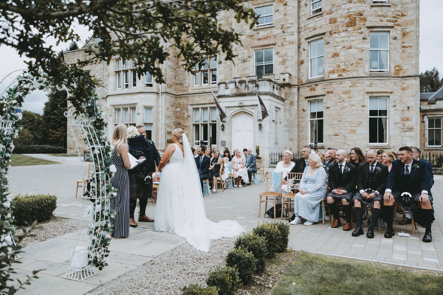 Elegant outdoor wedding ceremony at a grand stone mansion with decorative arch.