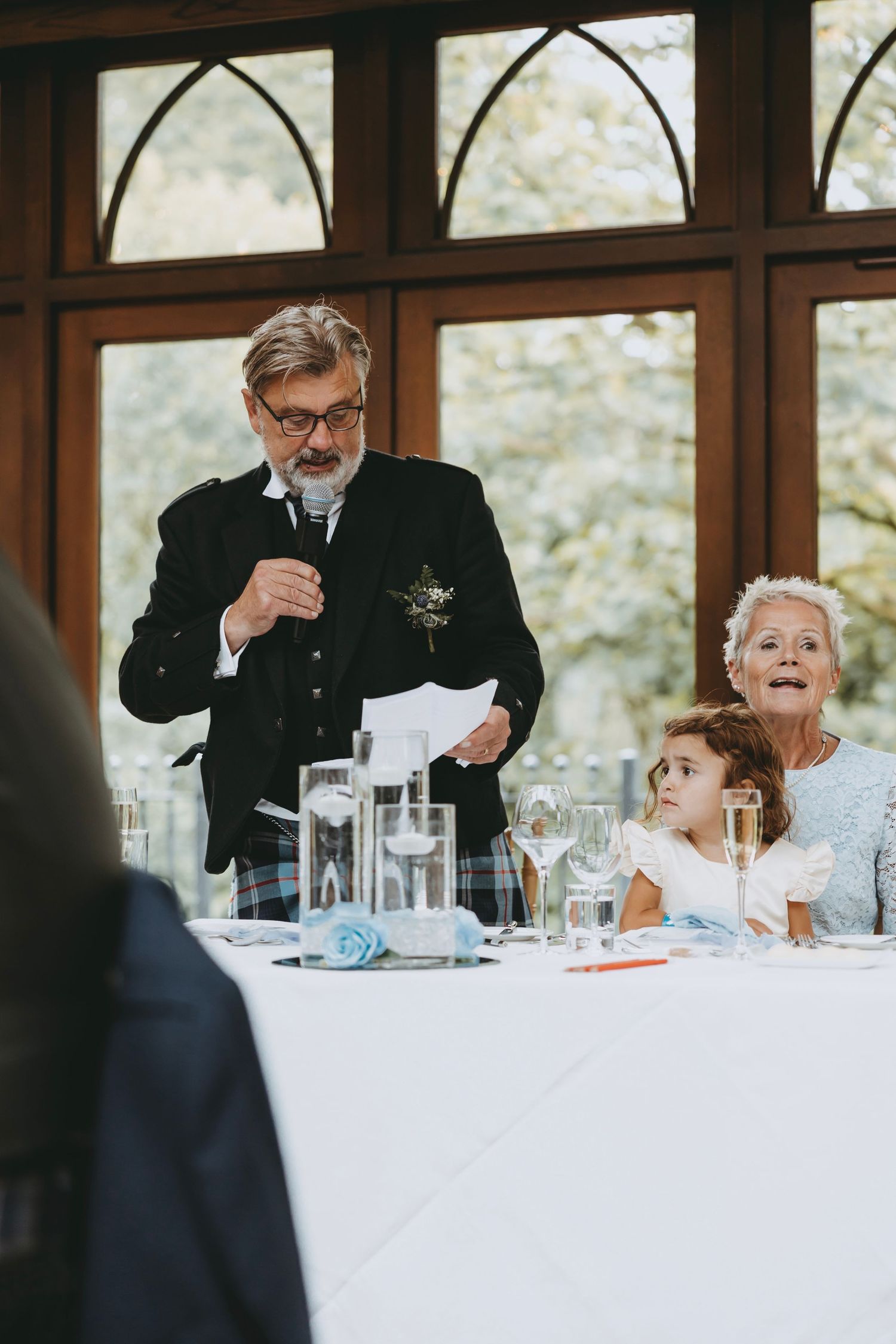 Wedding reception guests reacting during speeches at a formal dining table.