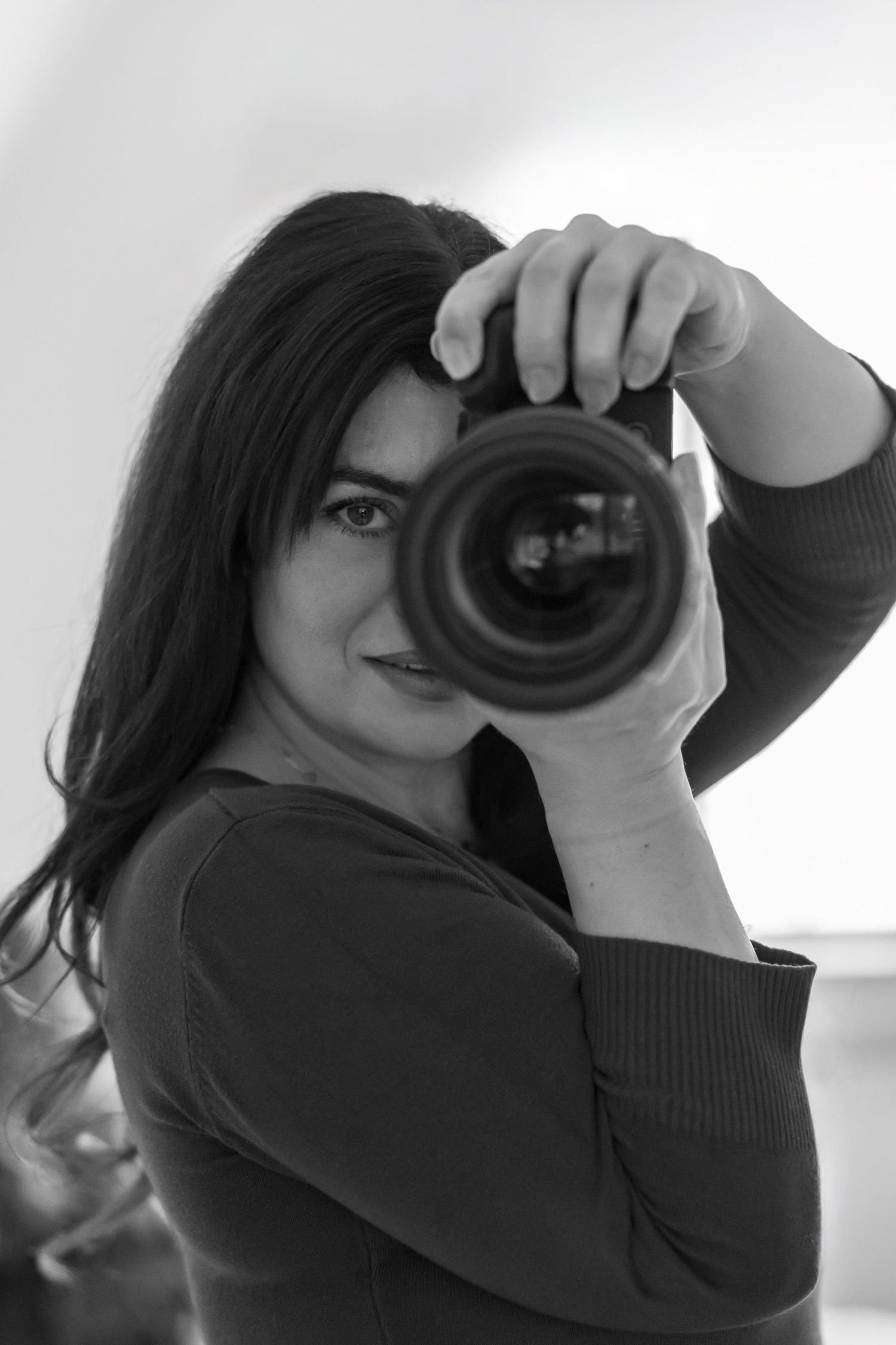 A photographer in black clothing holds up a professional camera with a large lens in a black and white portrait.