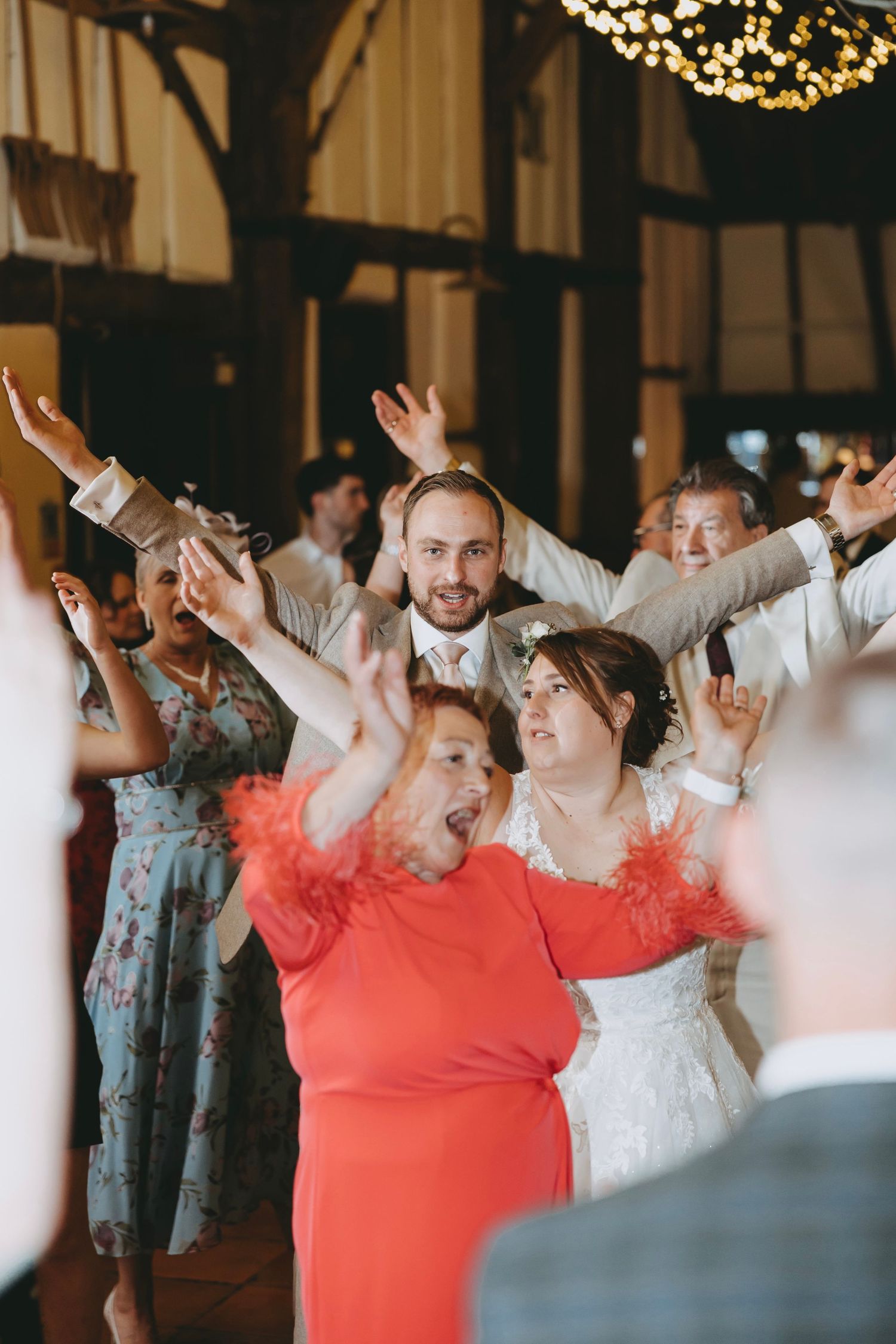 Wedding guests dancing joyfully at a rustic barn reception, bride in white dress and woman in red feather boa raising hands.