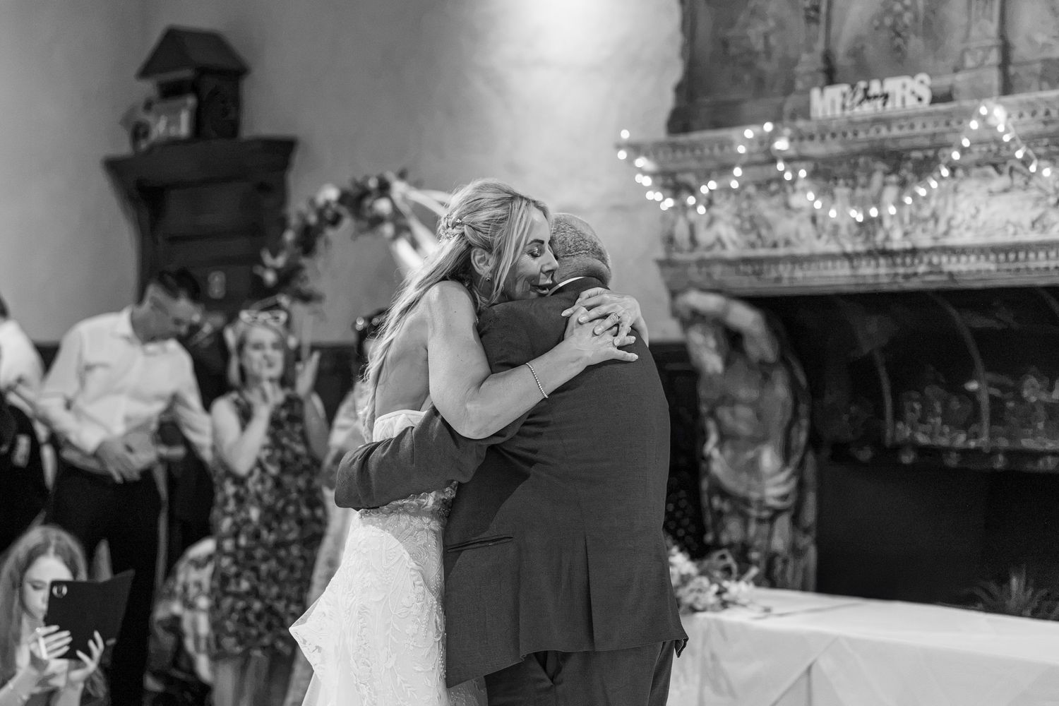 Black and white photo of a bride and groom sharing an emotional hug near a fireplace at their wedding reception.