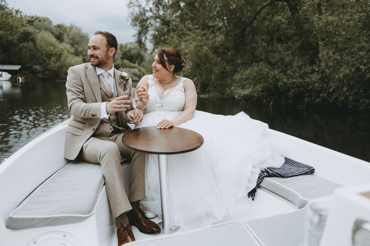 Bride and groom sitting on a white boat, toasting with champagne glasses on a scenic river surrounded by lush greenery.