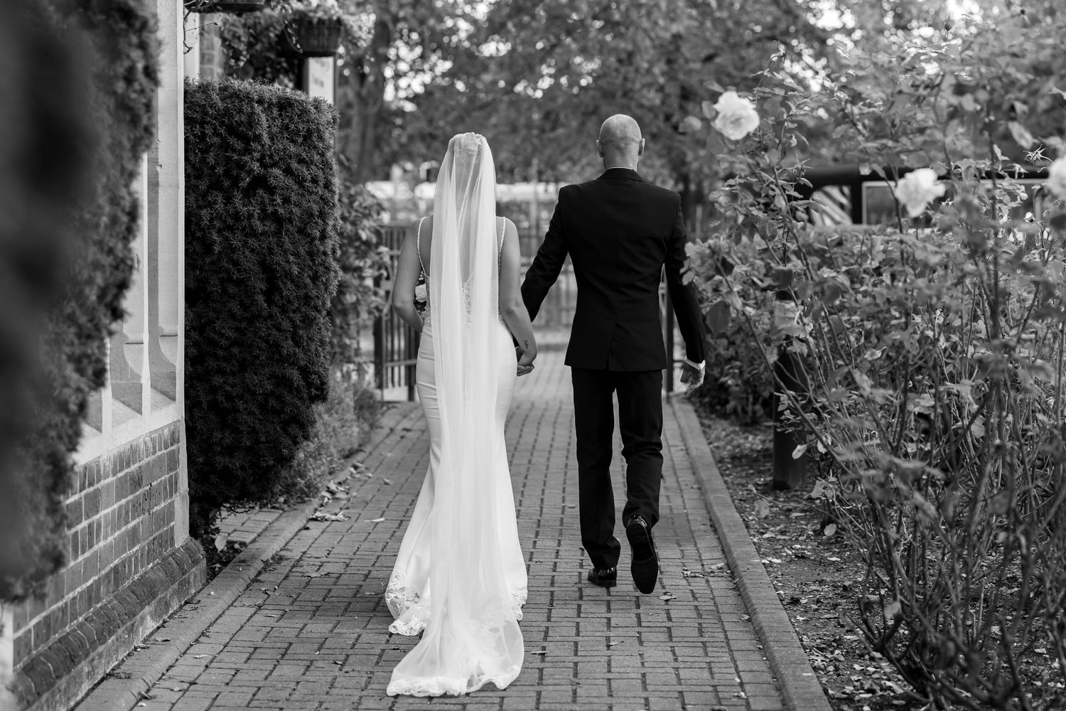 A bride and groom walk down a brick path together on their wedding day in black and white photography.