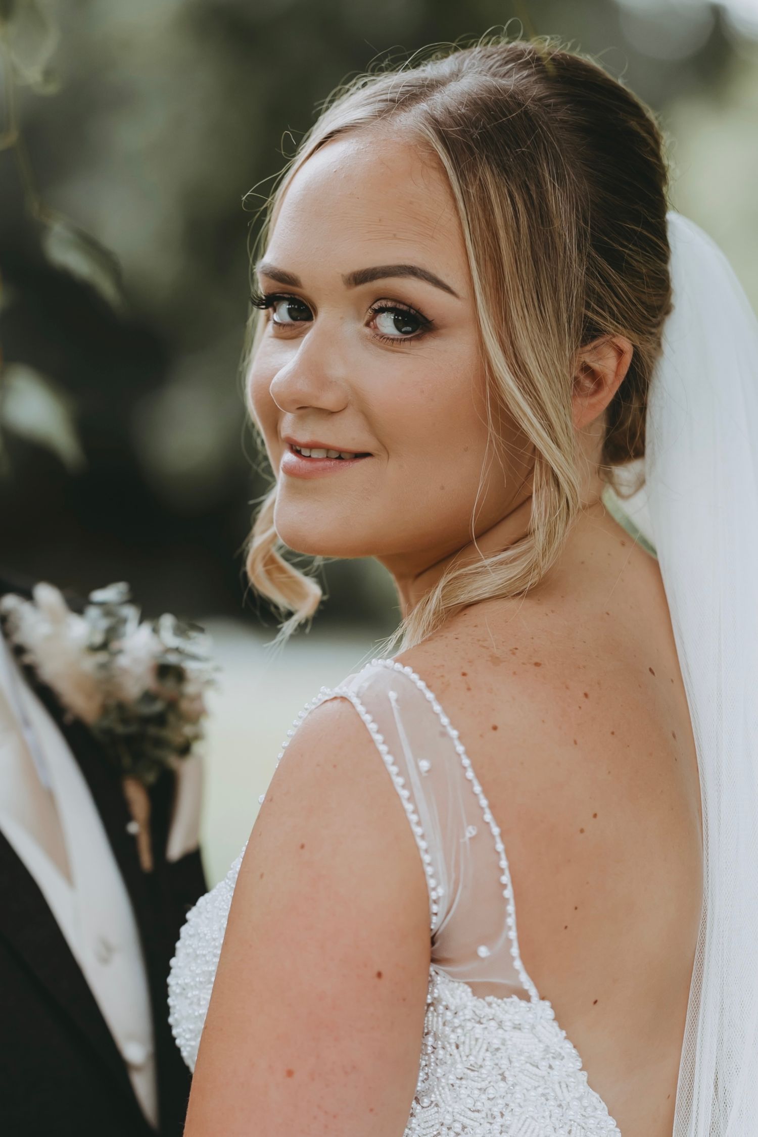 Bride wearing white lace dress and veil smiles over shoulder in outdoor portrait.