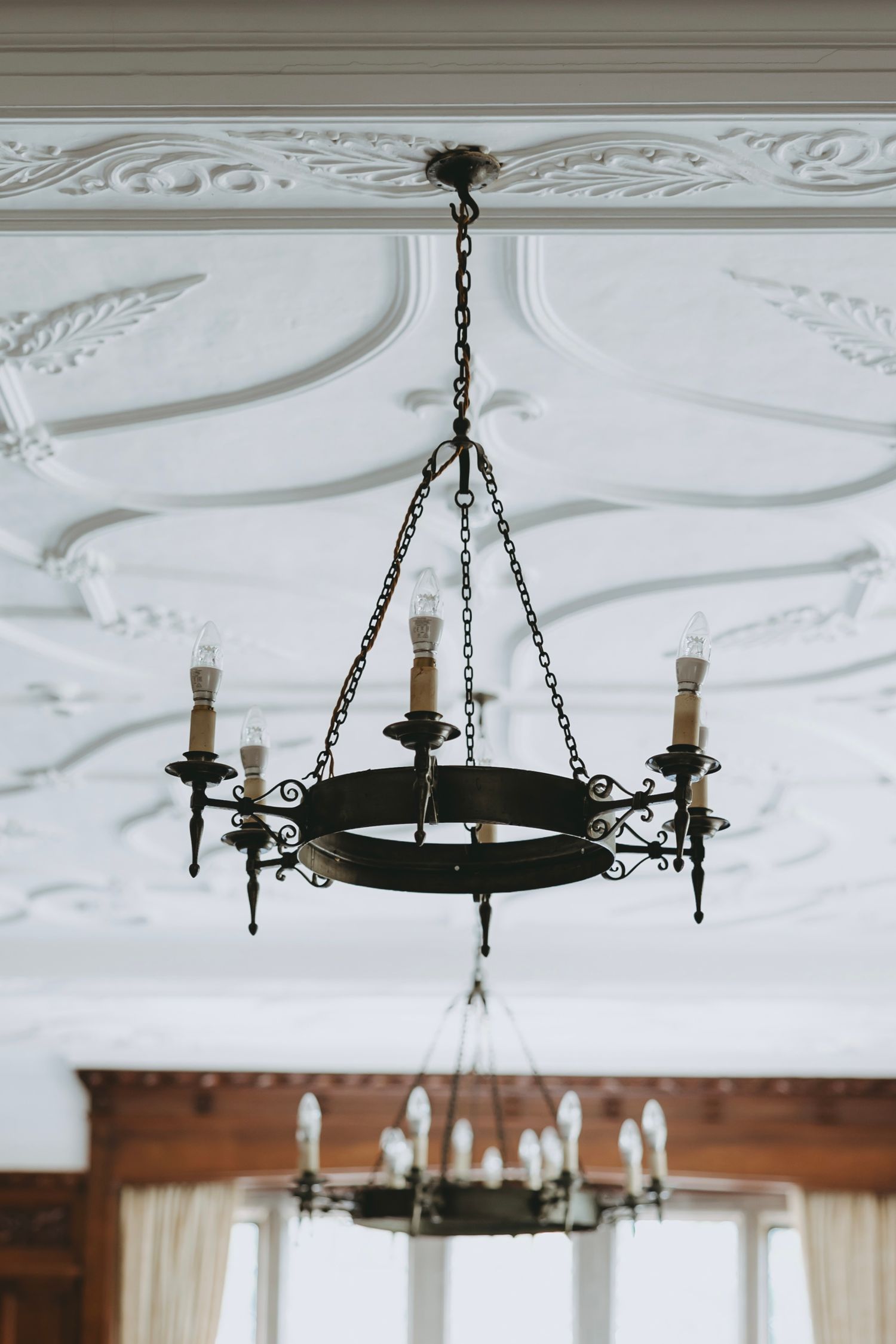 Vintage wrought iron chandelier with black metal ring and candle-style lights hangs from ornate white ceiling with moldings.