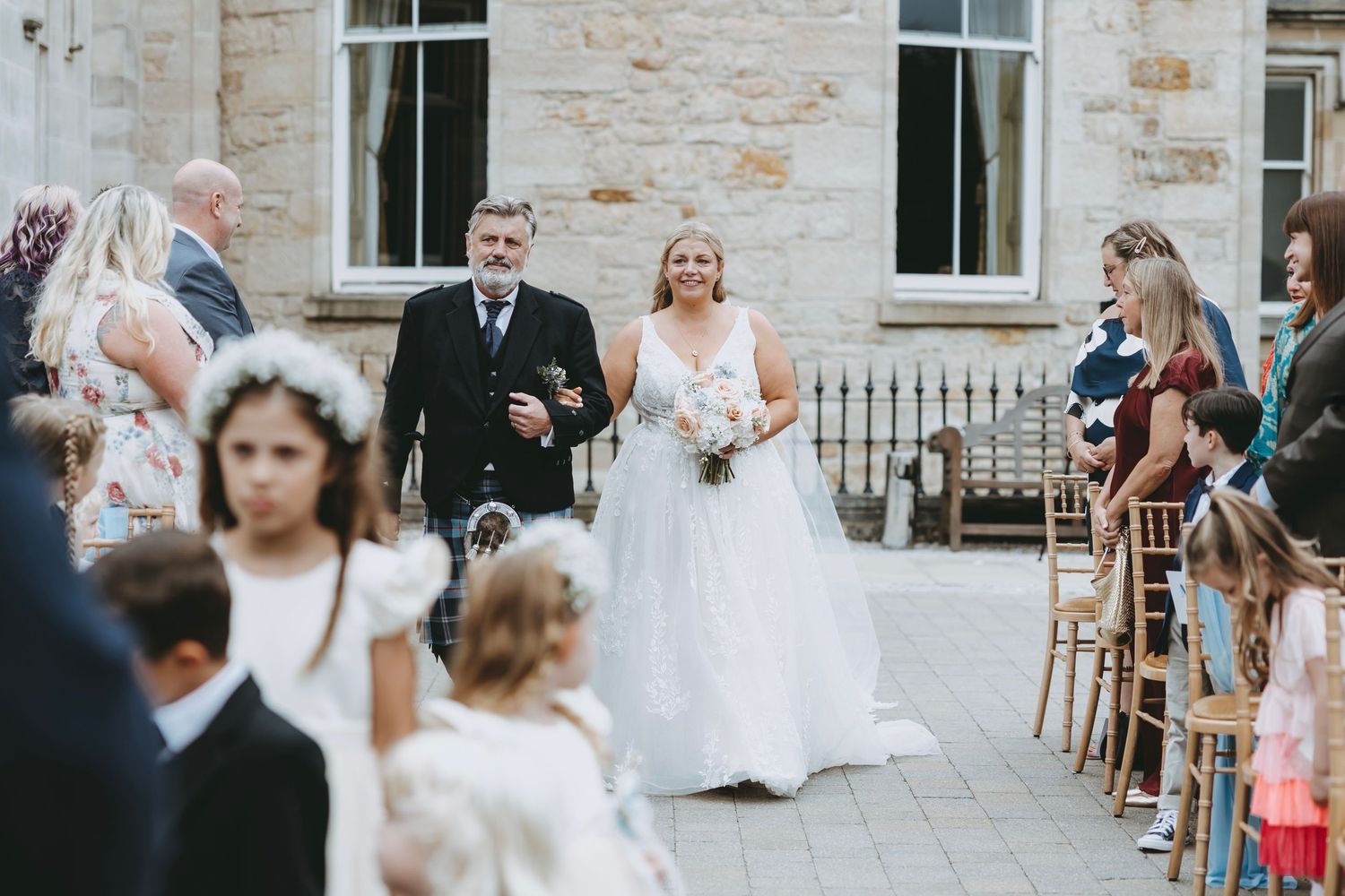 Wedding ceremony processional with guests seated on wooden chairs at stone venue.