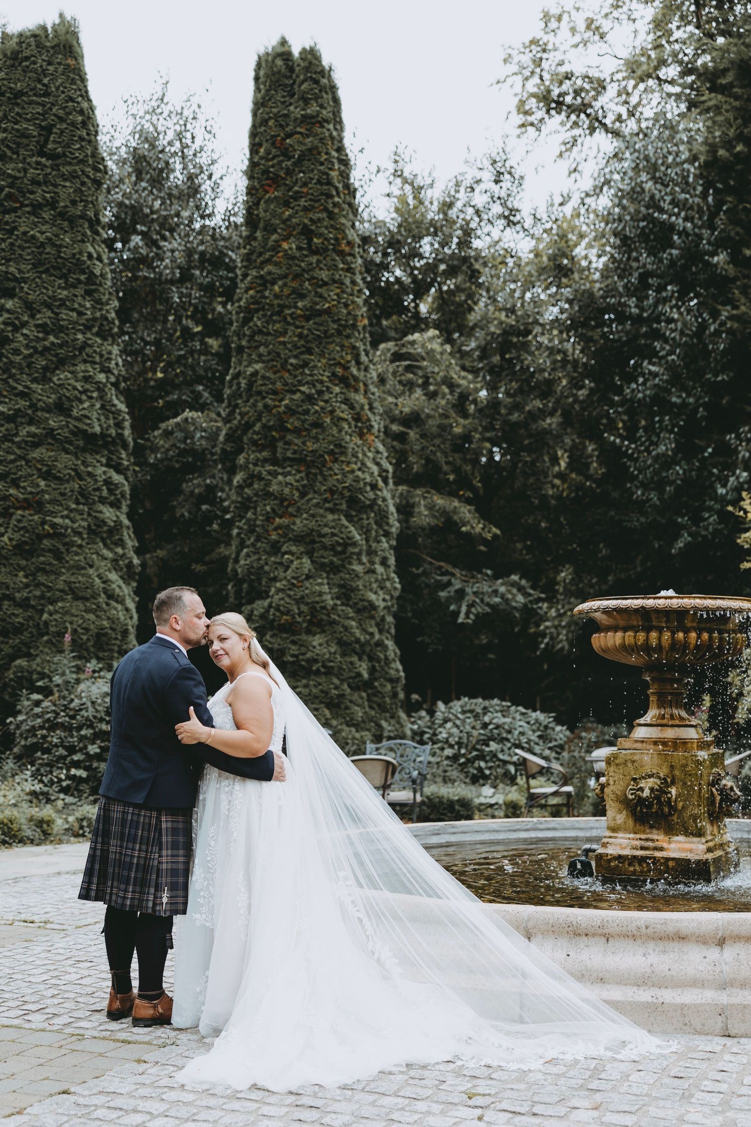 Wedding couple embracing near a decorative fountain in a garden setting with cypress trees at Crossbasket Castle, Scotland.