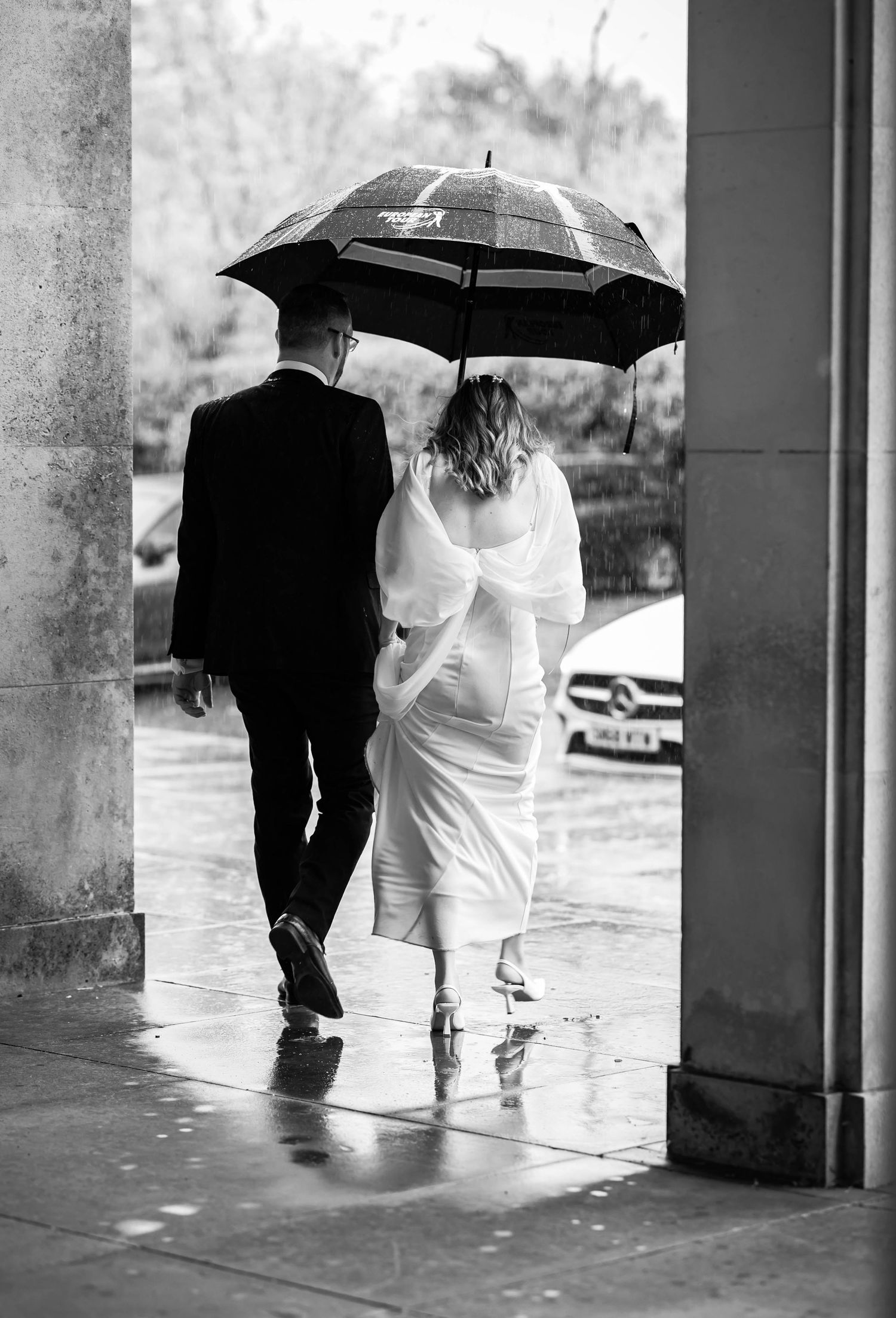 Black and white photo of couple with umbrellas walking through covered walkway in rain.