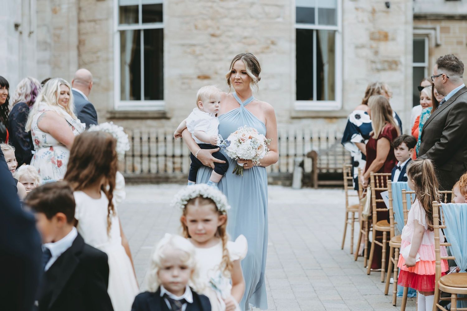Outdoor wedding scene with guests and children in formal attire at stone venue.