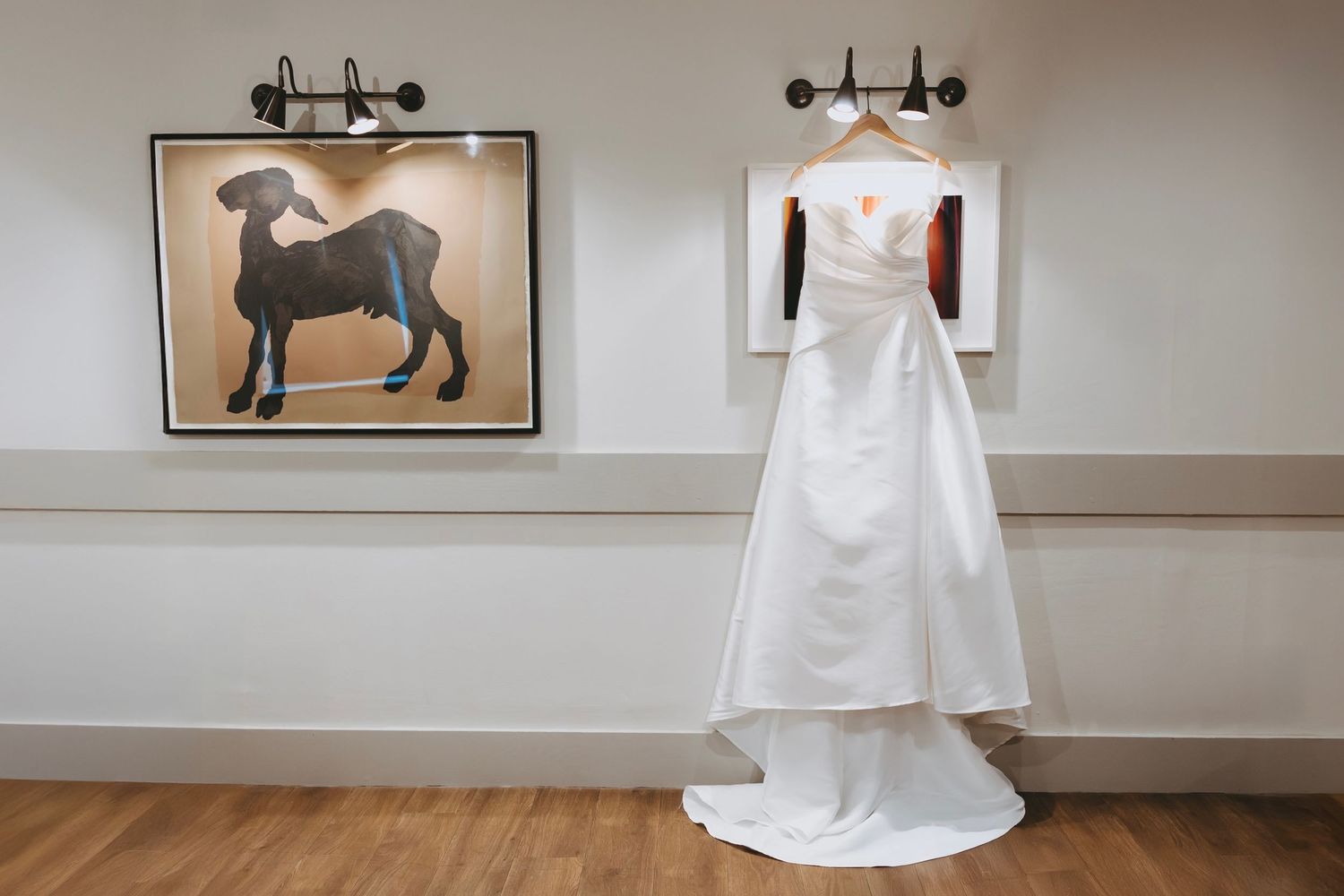 White wedding dress displayed on a hanger against neutral wall with decorative artwork.