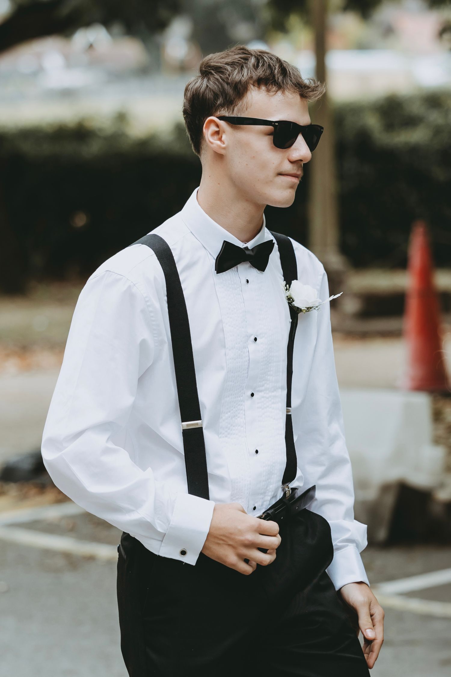 A well-dressed person in a white dress shirt, black bow tie, suspenders and sunglasses stands outdoors.