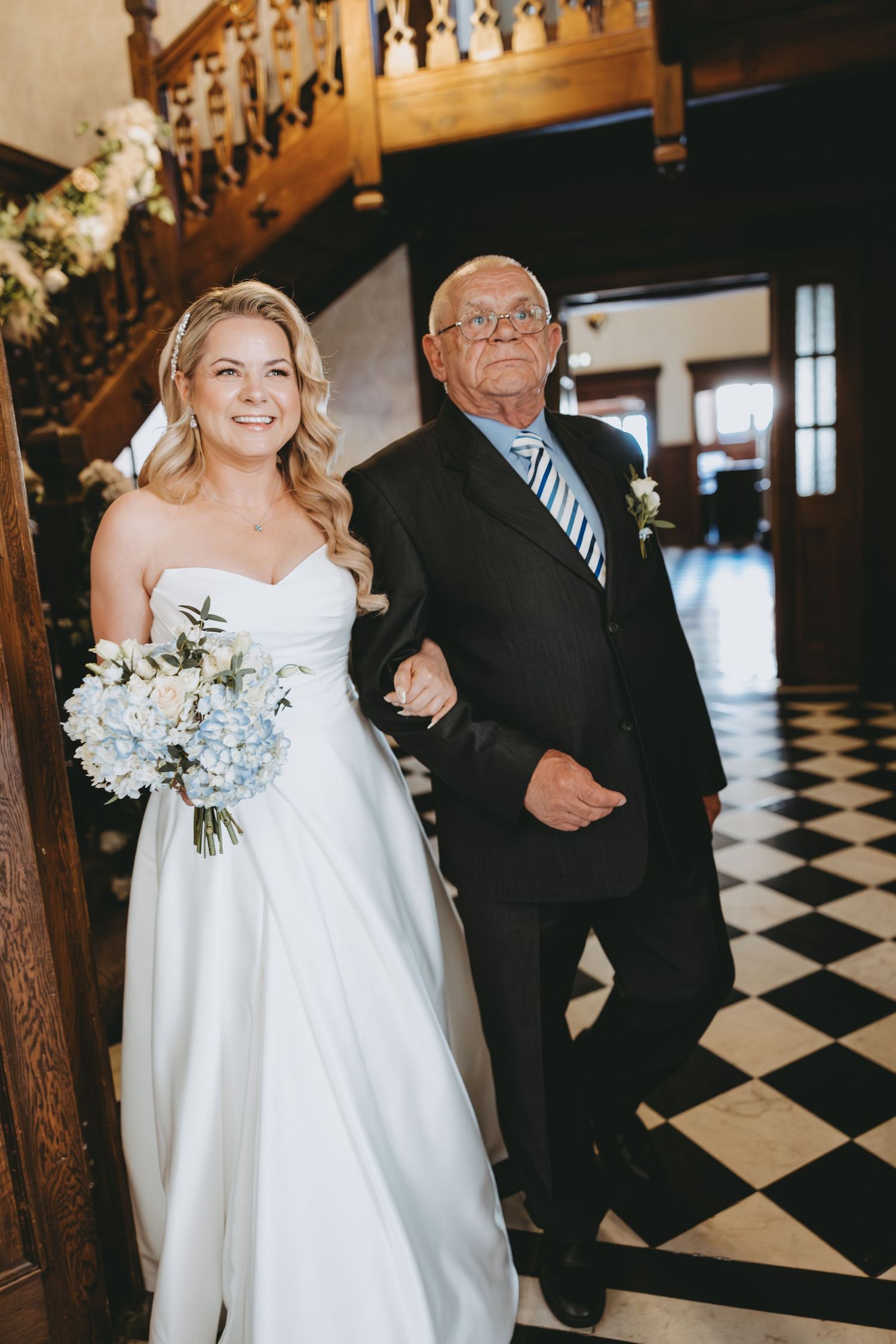 A bride in white dress walks down a black and white checkered floor with an escort.