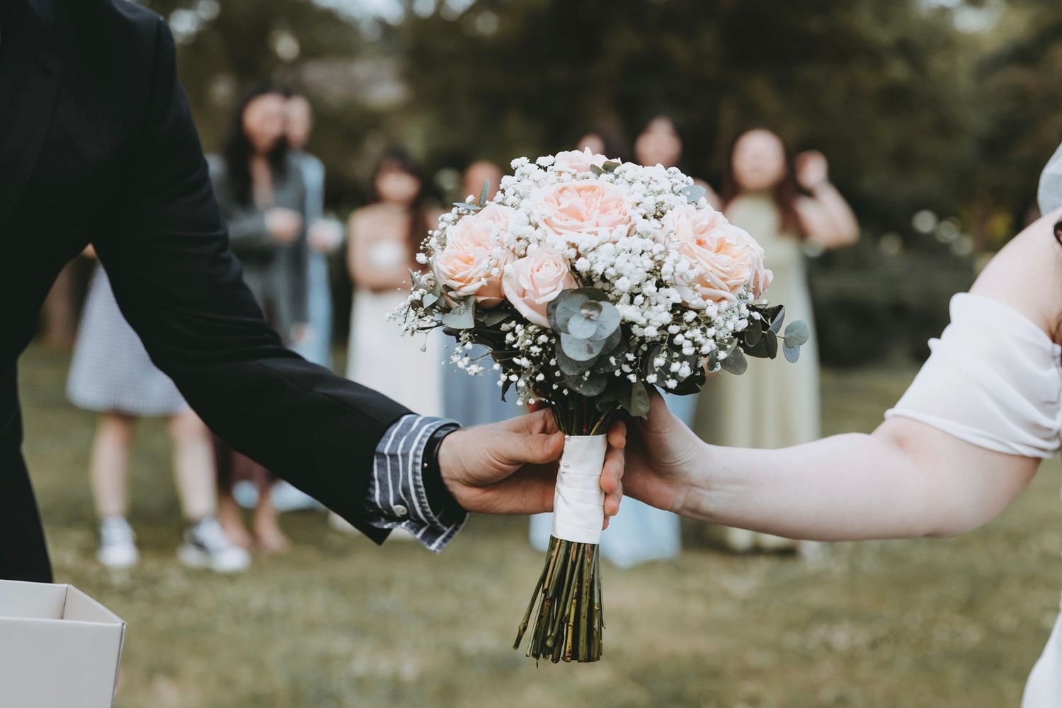 Groom presenting a pink rose and baby's breath bridal bouquet to bride at outdoor wedding ceremony.