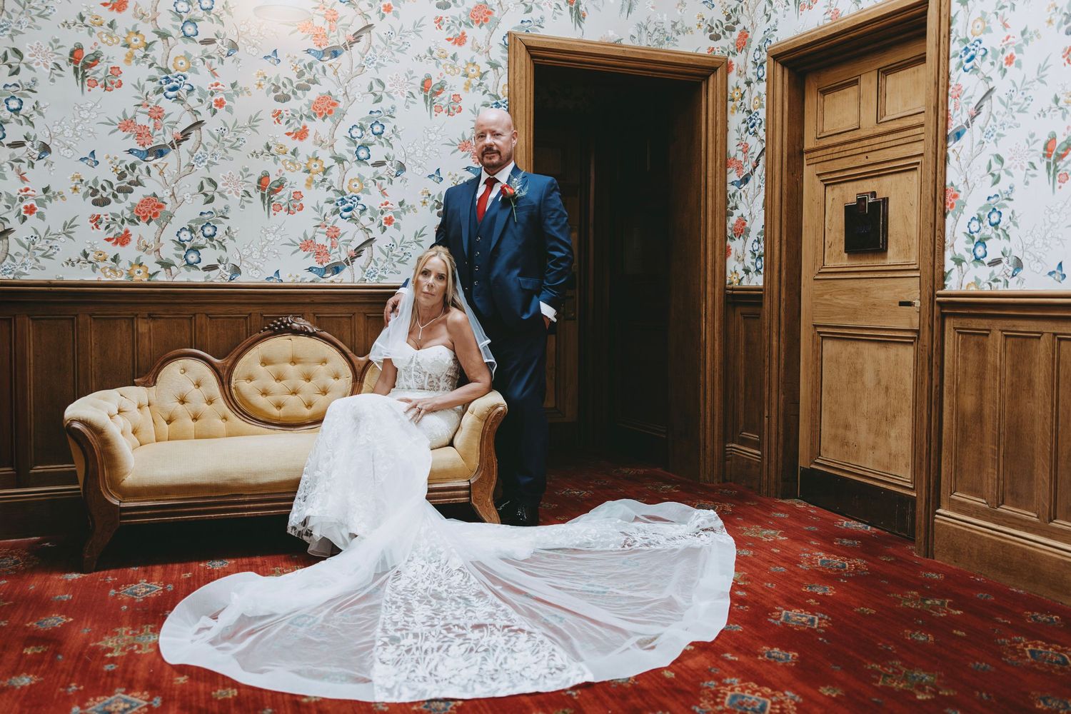 Bride in lace gown with long train sits on vintage sofa beside suited groom in ornate wallpapered wedding venue.