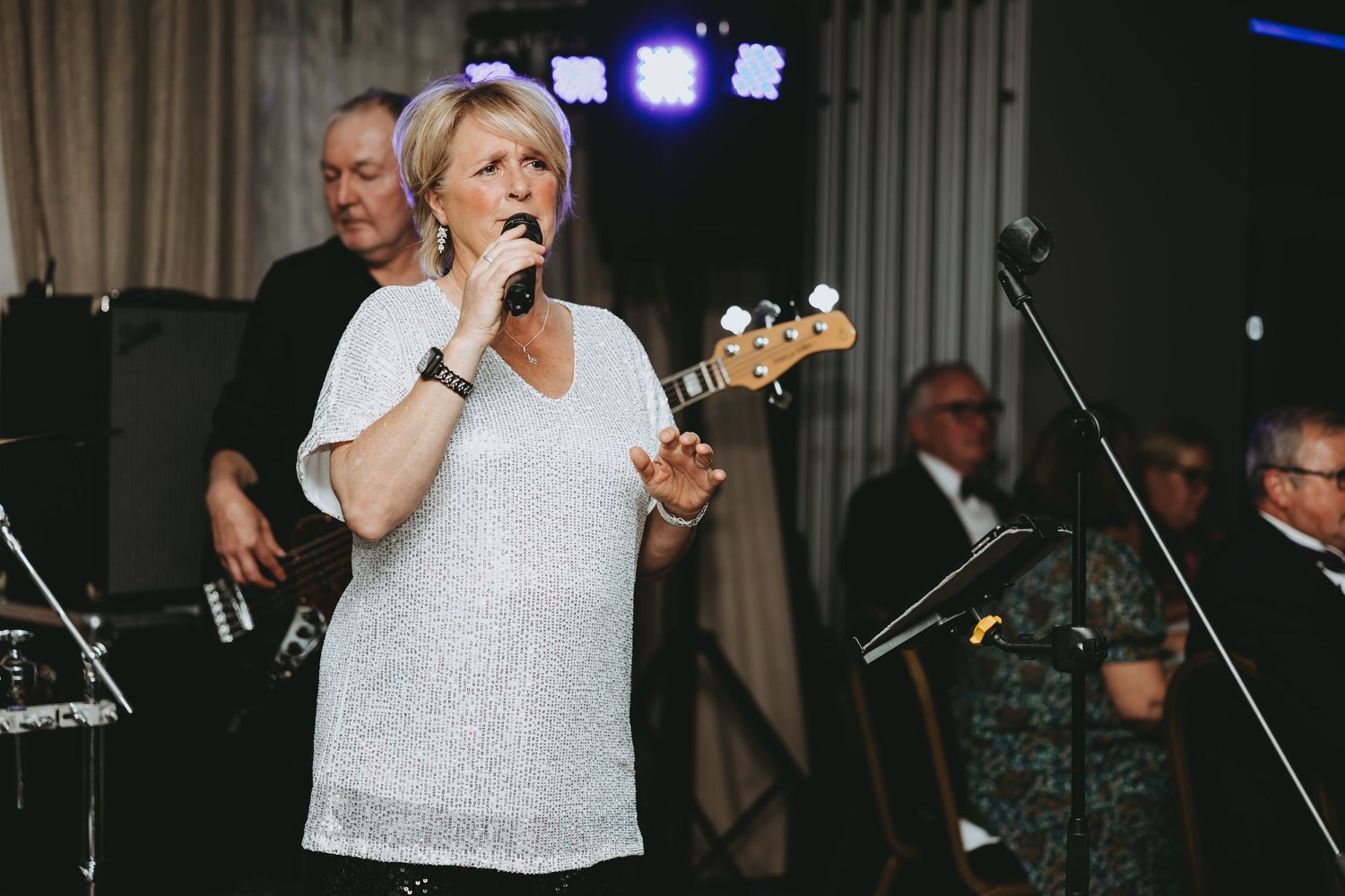 A performer in a white sequined top sings into a microphone on stage with band members visible in the background.