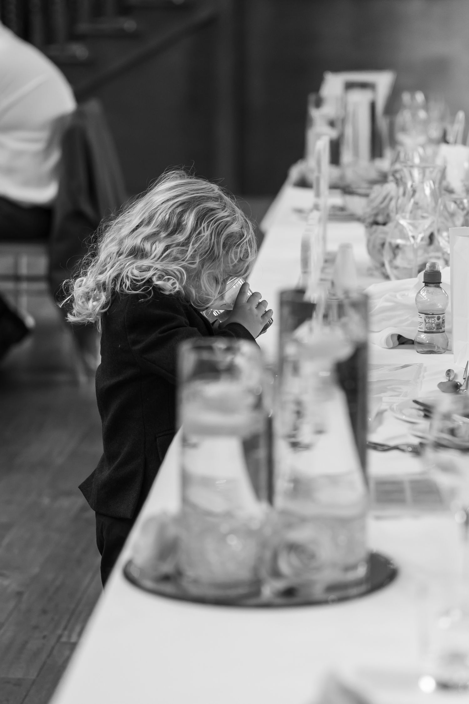 A black and white photo of a small child peeking over a table with glass bottles.