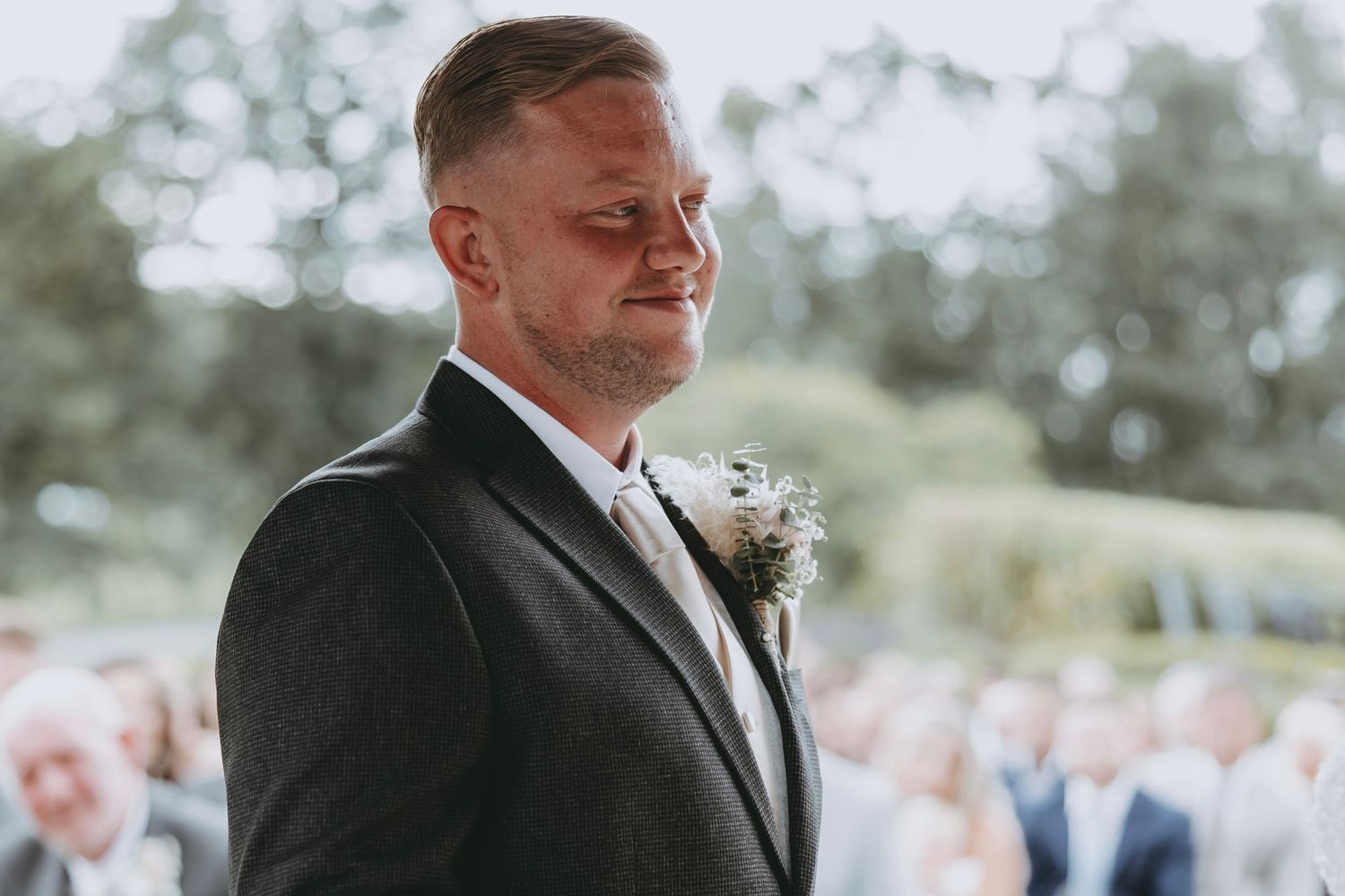 Groom in black suit listens intently during outdoor wedding ceremony.