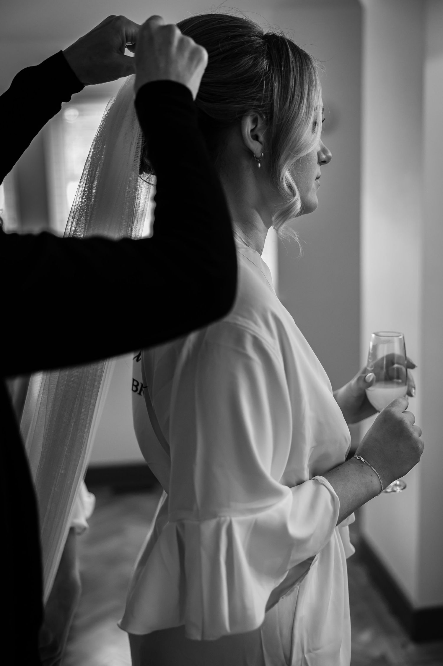 Black and white photo of wedding preparation with veil being adjusted while holding a drink.