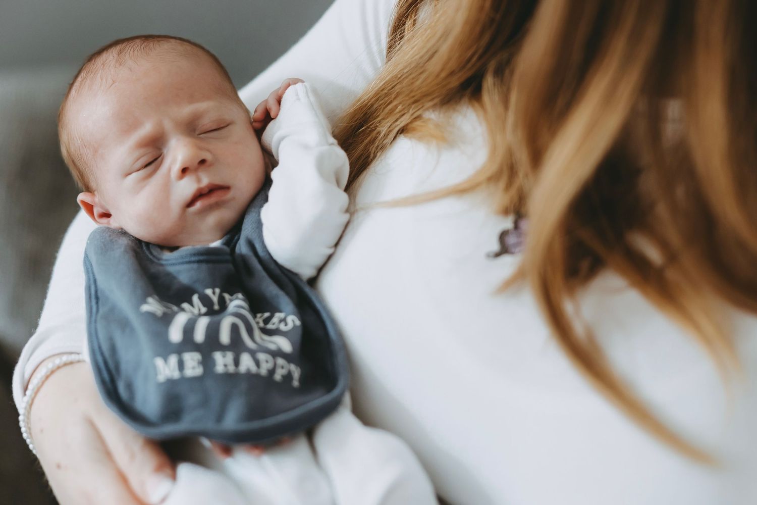 A sleeping baby wearing a gray onesie rests peacefully while being held in someone's arms.