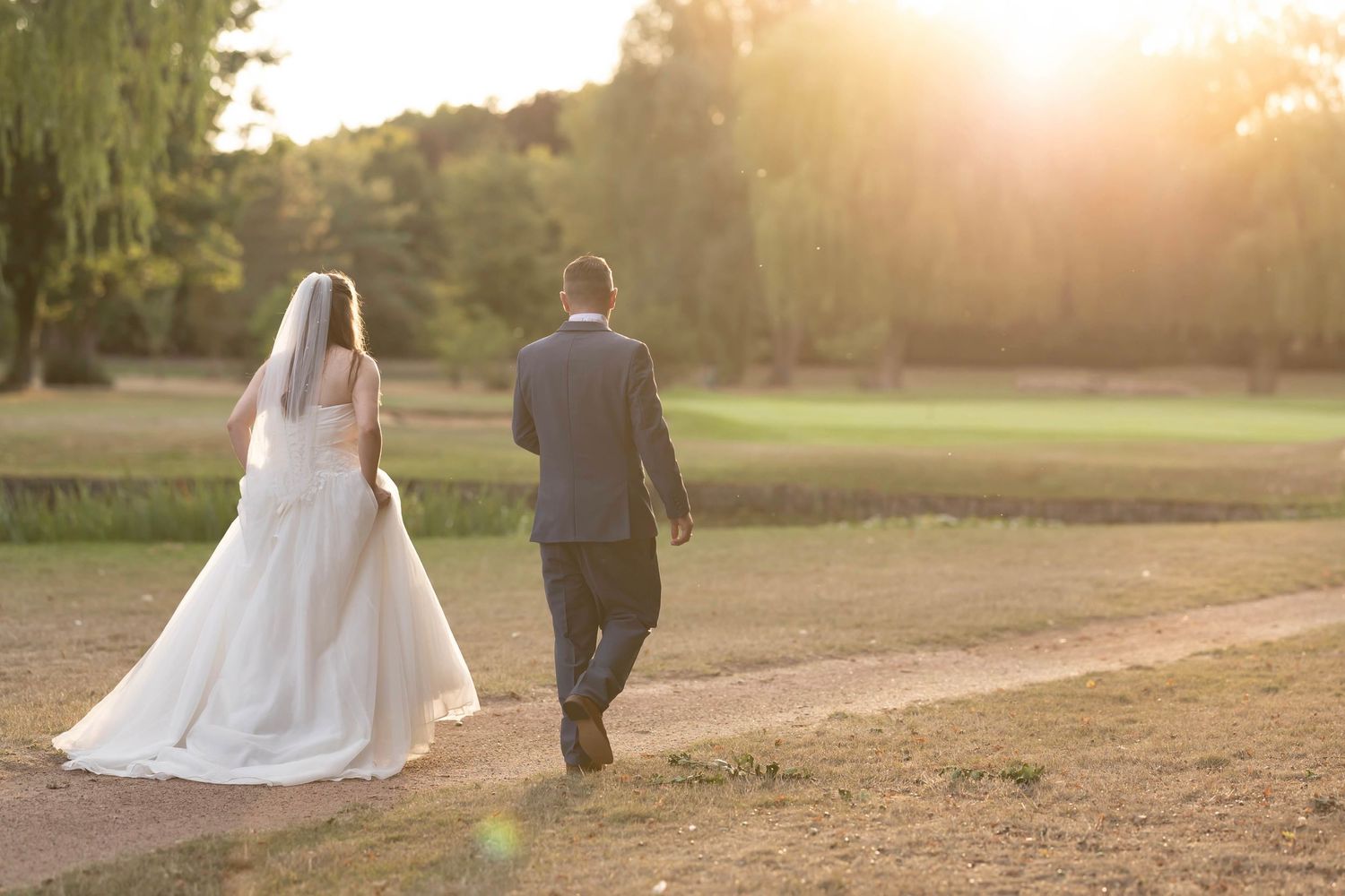Silhouetted couple walks hand in hand through a sunlit field at golden hour as their wedding attire glows in warm light.