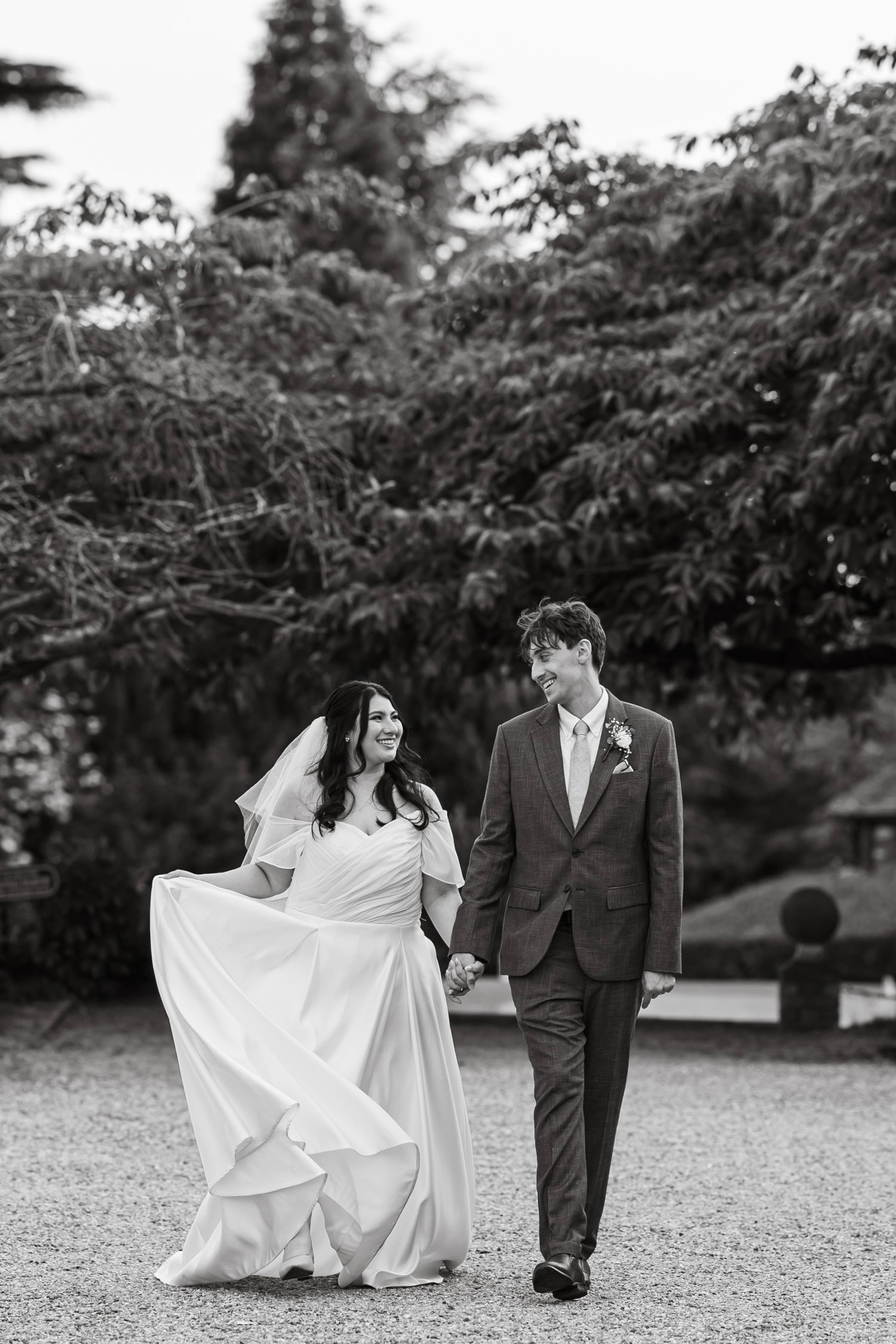 Black and white photo of a bride and groom walking hand in hand through garden grounds on their wedding day.
