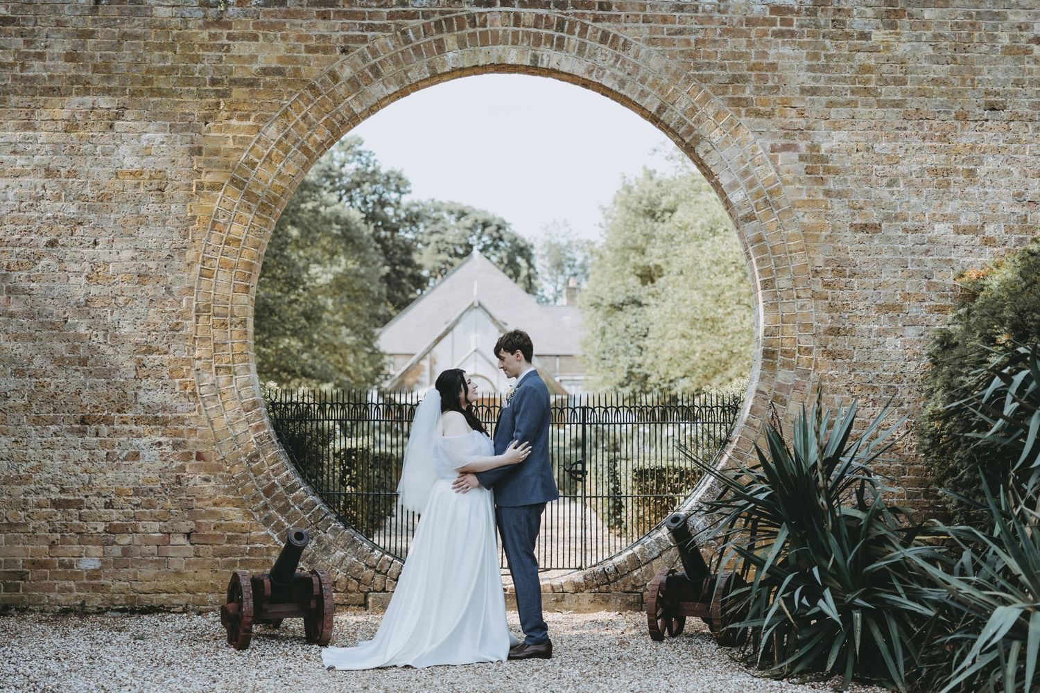 Bride and groom share a romantic moment framed by a circular stone arch in a garden wedding setting.
