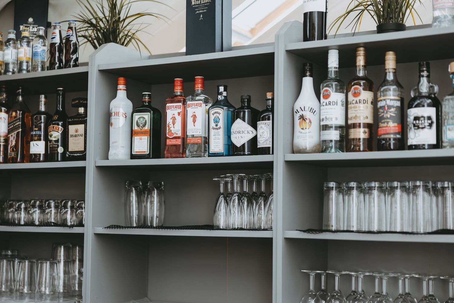 Gray bar shelves stocked with assorted liquor bottles and rows of empty glasses in a modern bar setting.